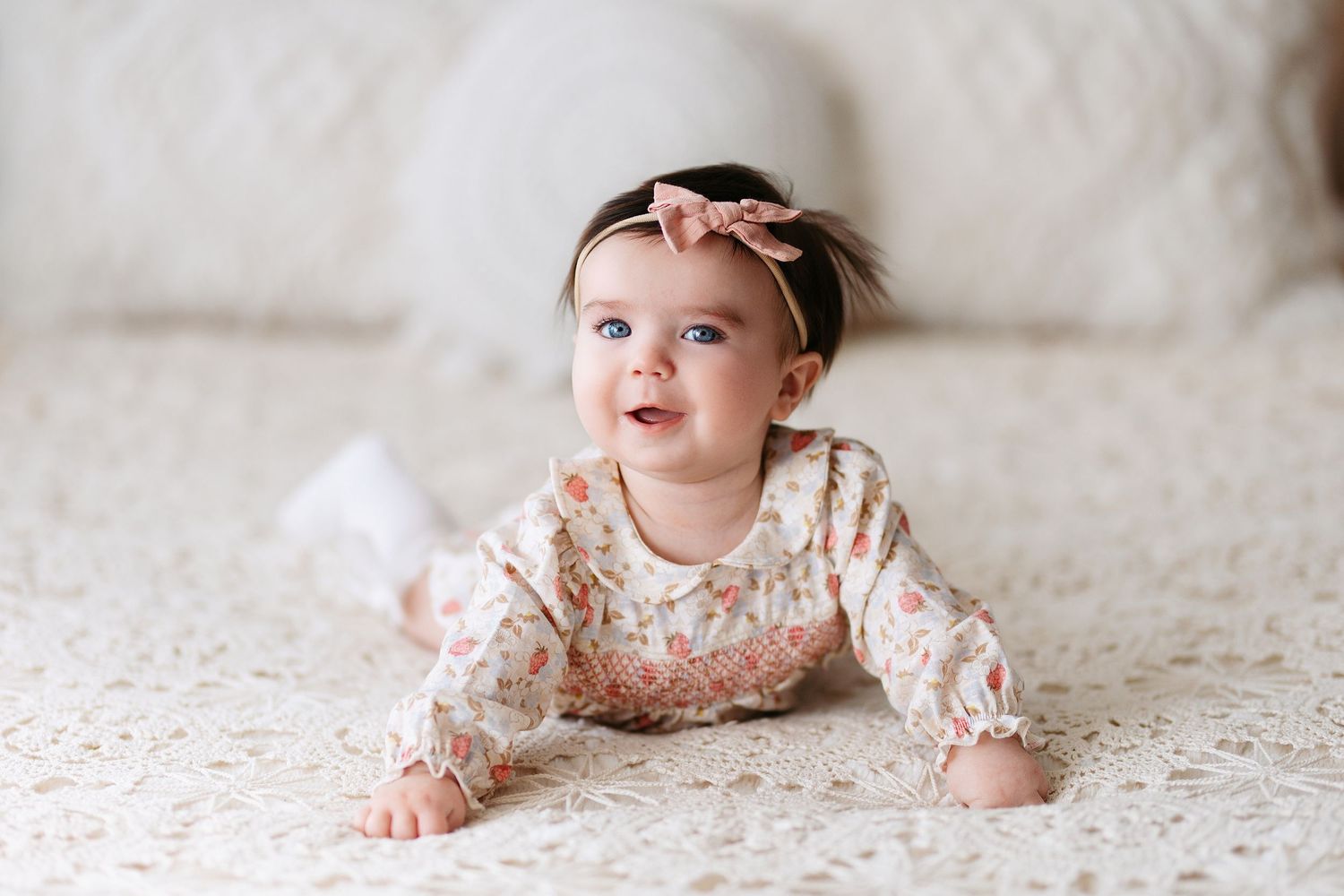 Baby in a floral dress lying on white textured bedding with a joyful expression.