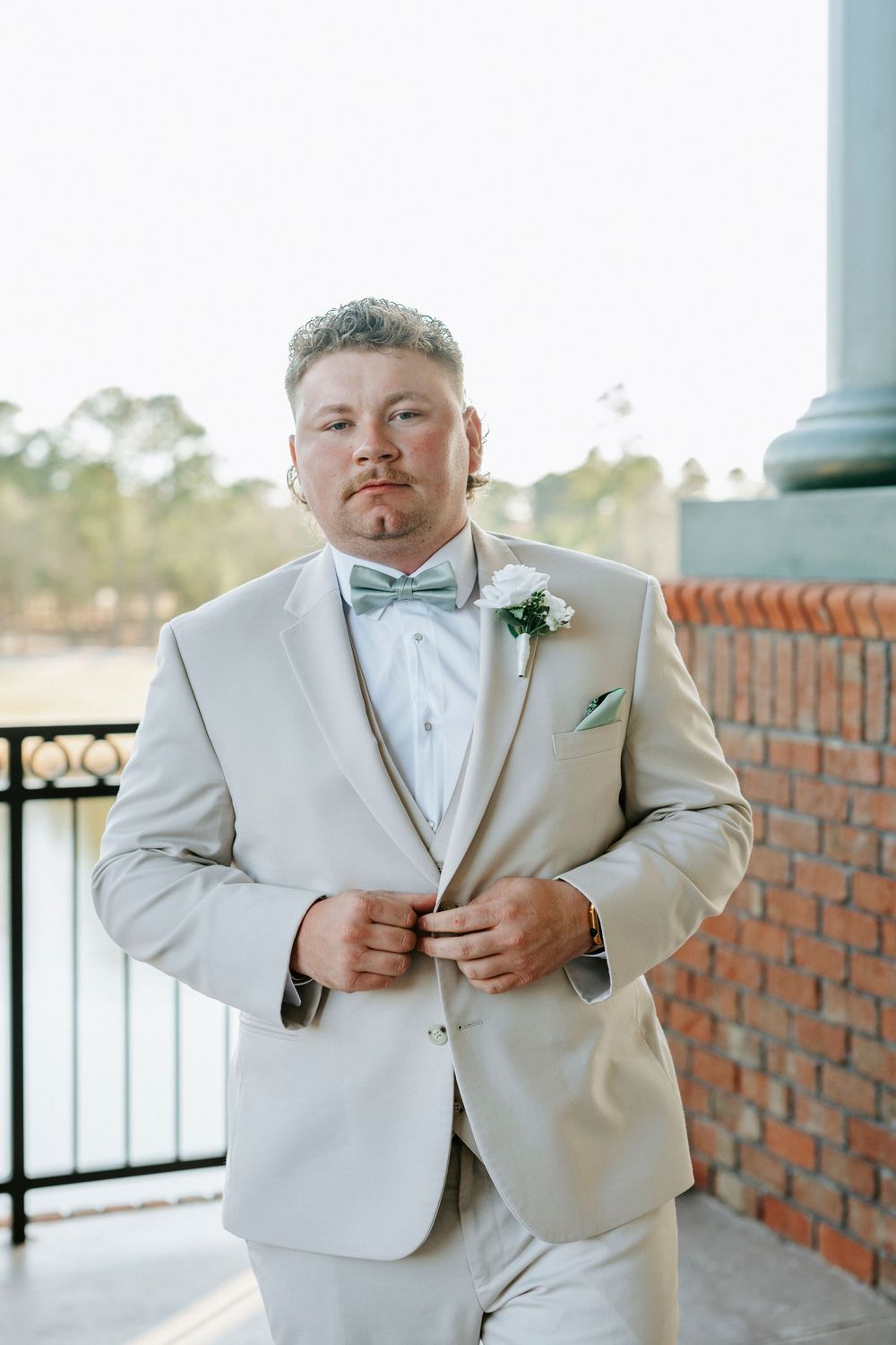 A groom in a light beige suit with mint bow tie and boutonniere poses against a brick wall.