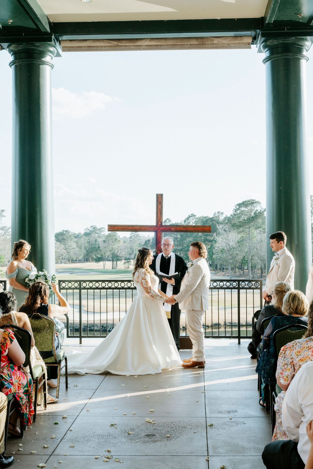 Wedding ceremony taking place under a covered outdoor pavilion with white columns and guests seated in rows.