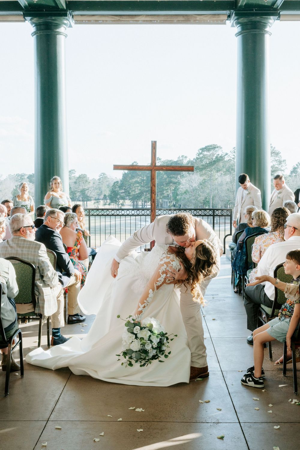Wedding party members sit at long tables under a covered outdoor venue during a reception celebration.