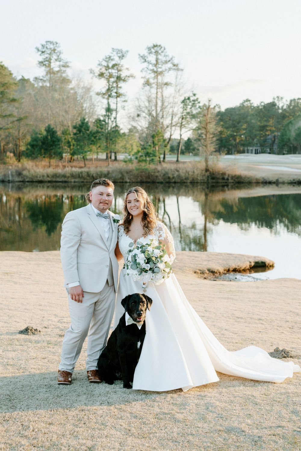 A couple poses with their dog by a serene lake with tall pine trees in the background.