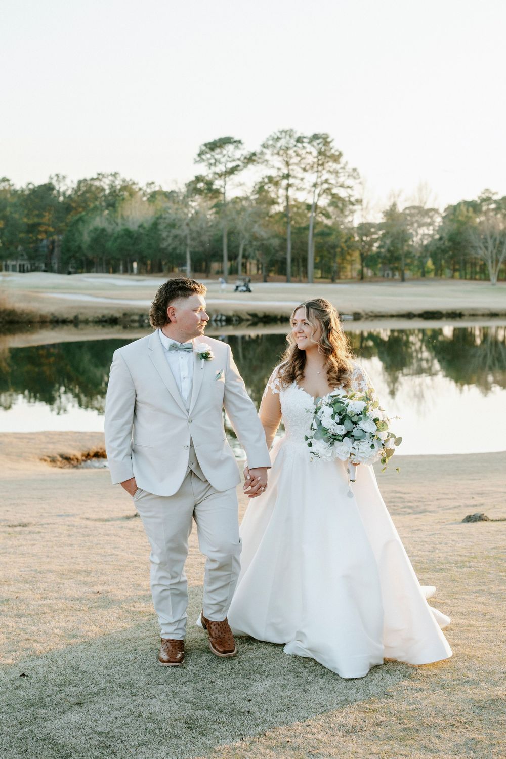A couple in wedding attire sits together by a serene lake during golden hour photography session.