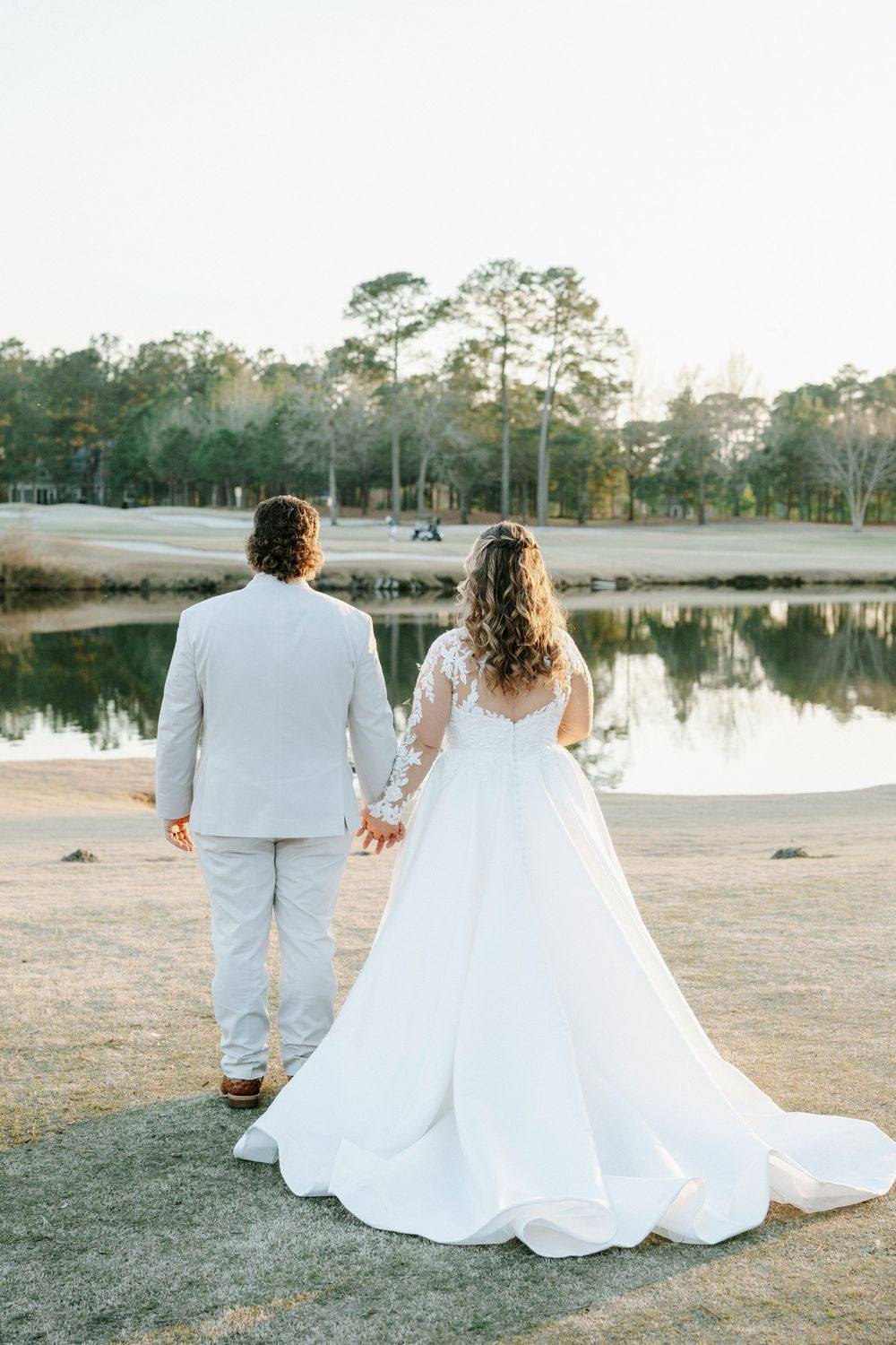 A newlywed couple sits together on grass near a serene lake at sunset, with the bride in a flowing white gown.
