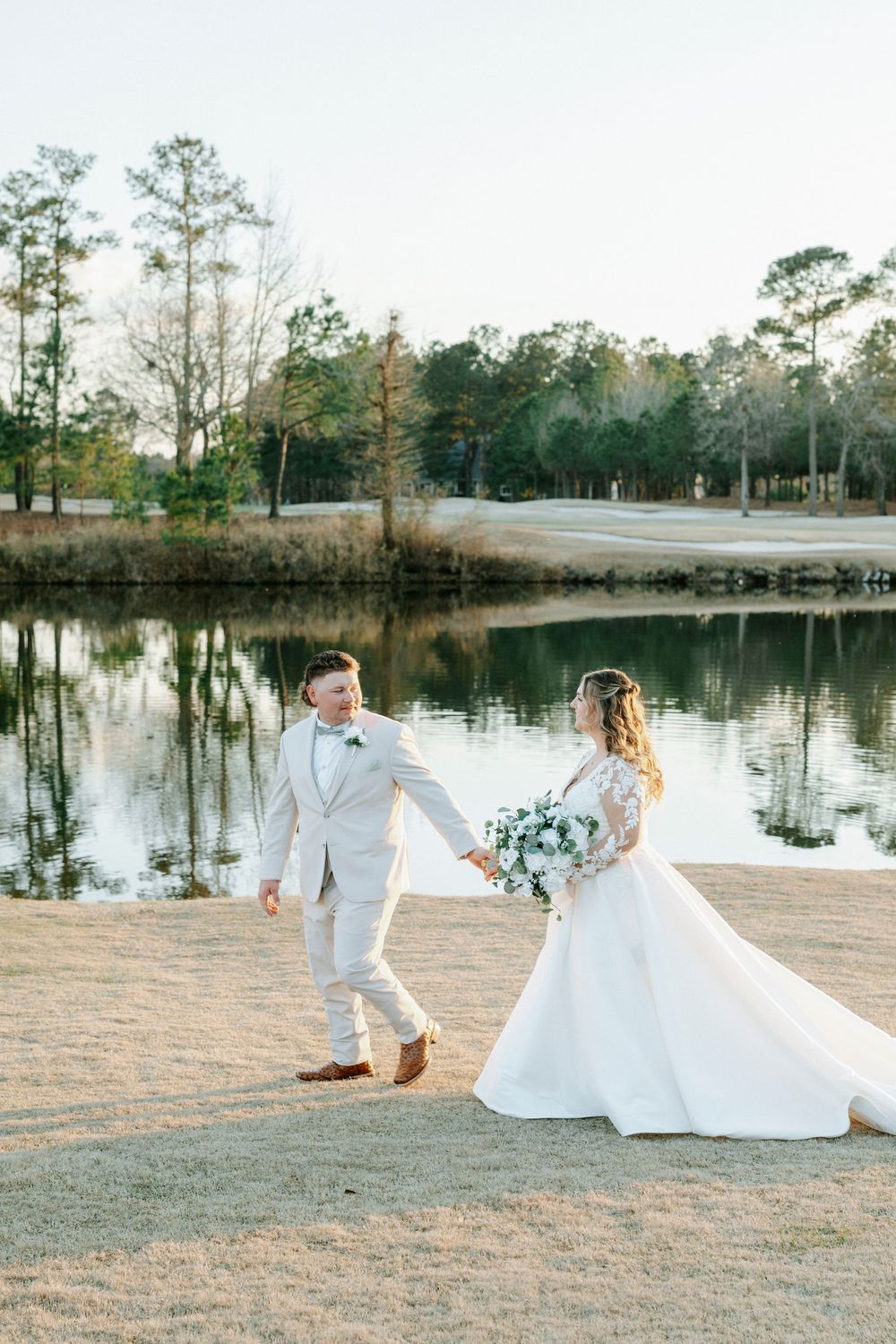 A couple in white wedding attire dances together by a serene lake at sunset with tall pine trees in the background.