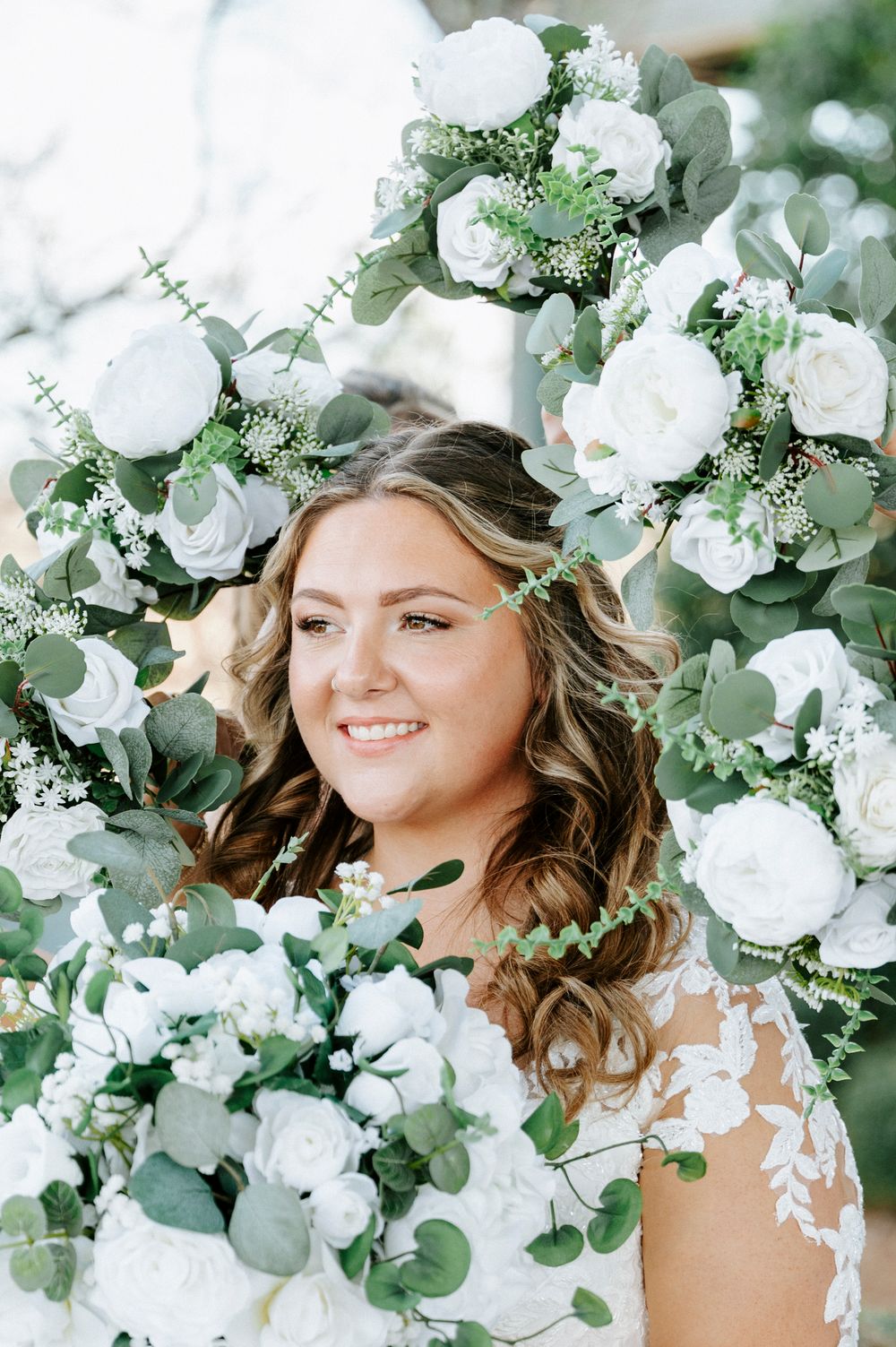 Ethereal bridal portrait surrounded by white roses and eucalyptus greenery in a dreamy garden setting.