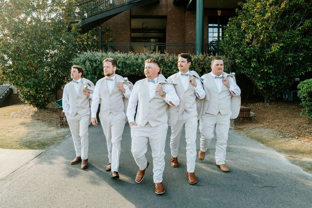 A group of groomsmen in light gray suits walk together on a stone pathway during a wedding celebration.