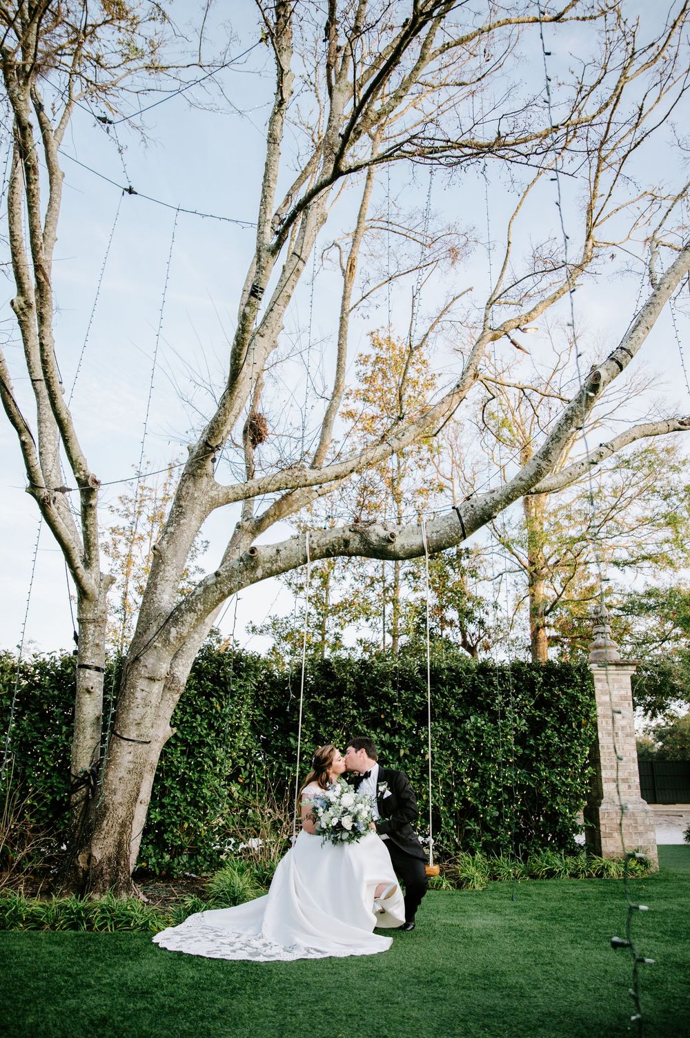 Couple enjoys romantic moments on tree-lined lawn with white wedding dress and greenery in background.