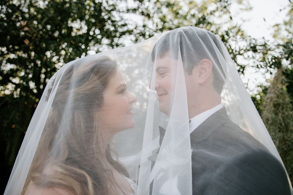 Romantic newlyweds share tender moments under a white wedding veil in a garden setting.