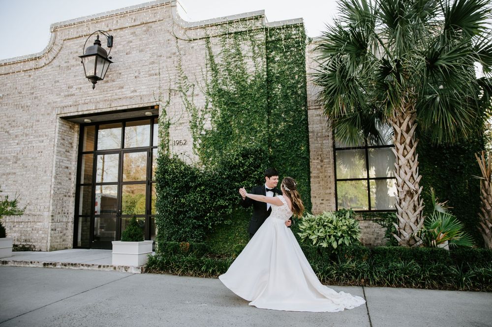 A bride in a flowing white gown twirls in front of a stone building with palm trees and climbing ivy.