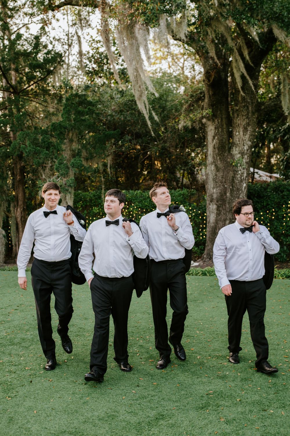 Four groomsmen in matching black and white attire walk together on a green lawn with moss-draped trees in the background.