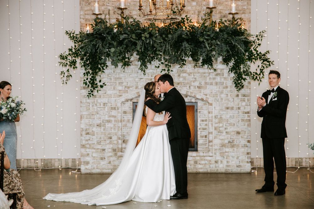 A romantic wedding ceremony moment in front of a whitewashed brick wall decorated with cascading greenery and candles.