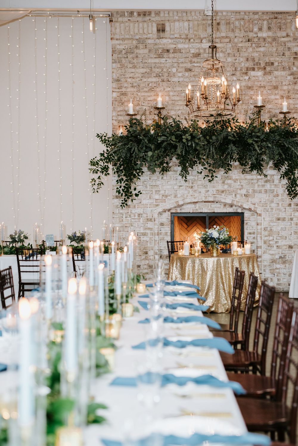 Elegant wedding reception tables with white linens and greenery decor against exposed brick walls in a rustic venue.