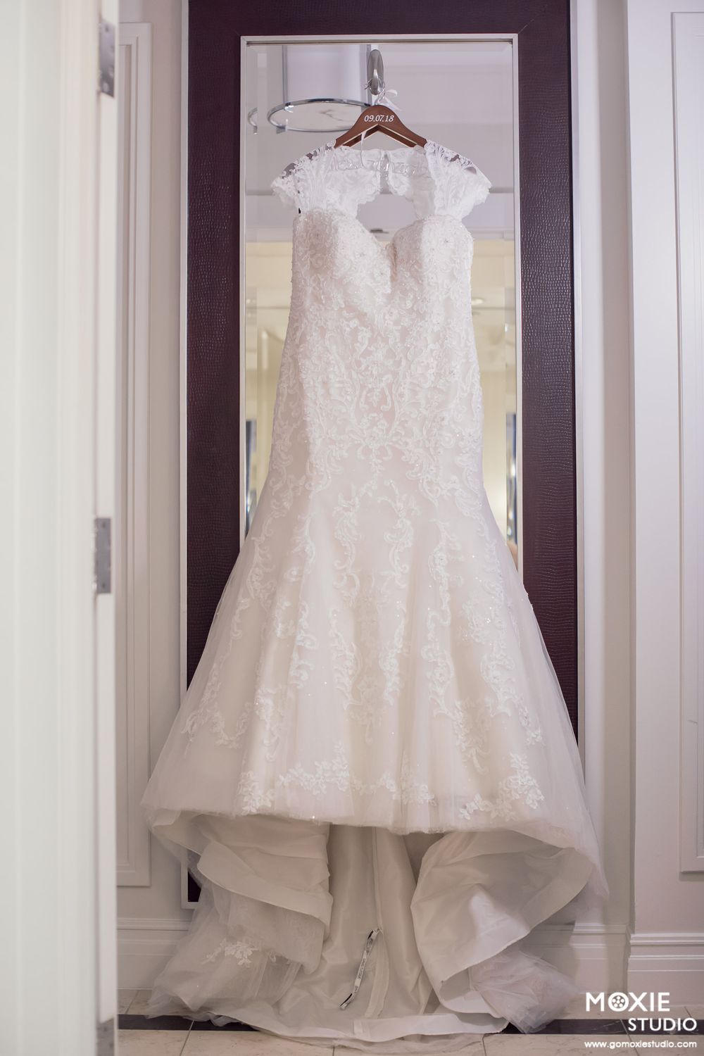 White wedding dress displayed on a hanger against a light-colored wall with multiple reflections in mirrors.