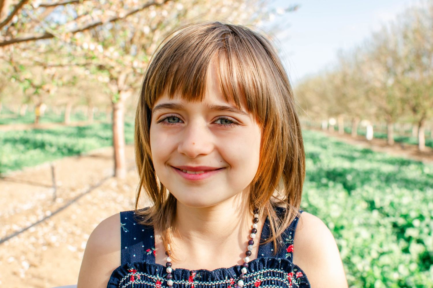 A person in a blue dress smiles while standing in a sunny field with white flowers and trees in the background.