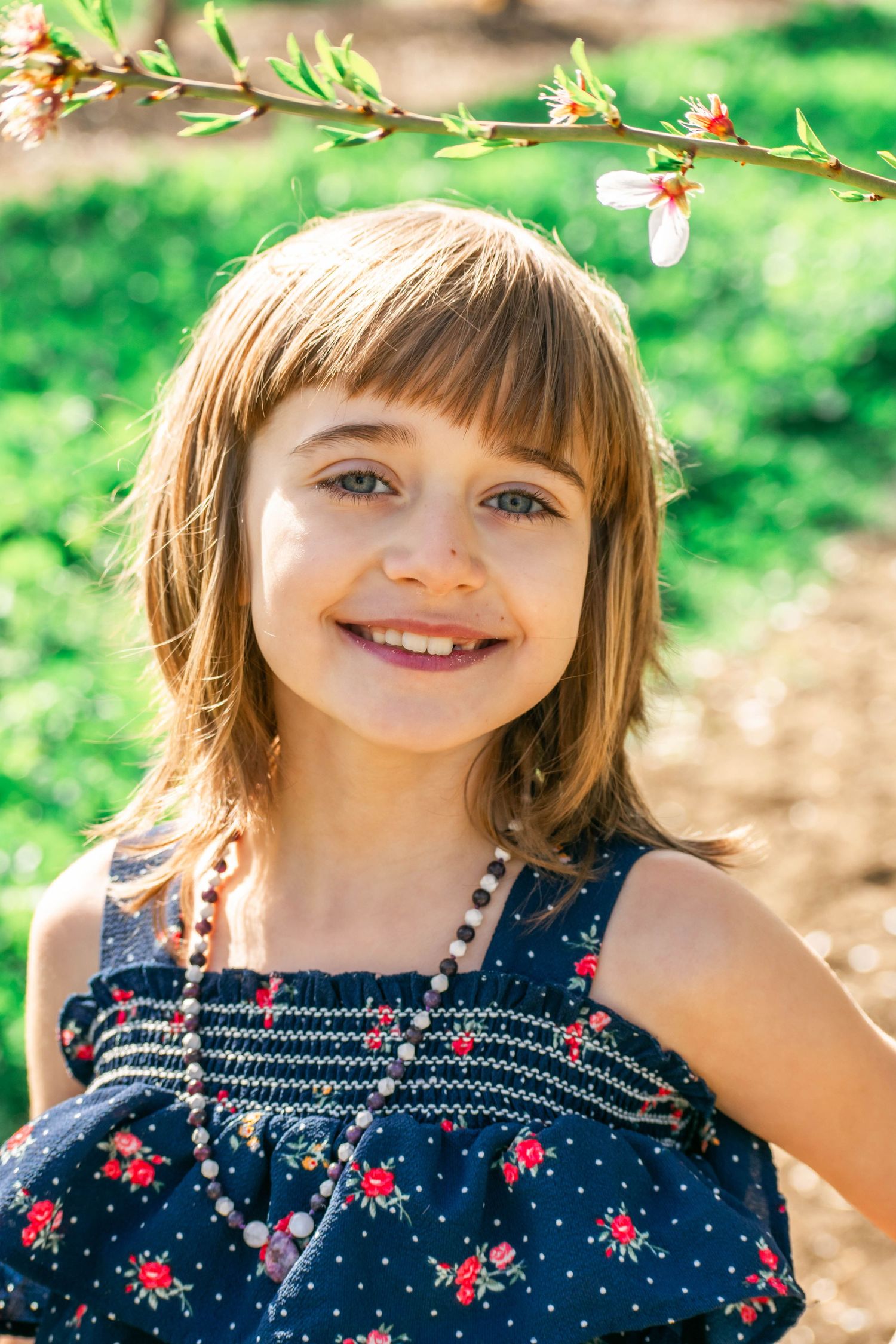 A child in a navy polka dot dress with red flowers smiles brightly in a sunny outdoor garden setting.