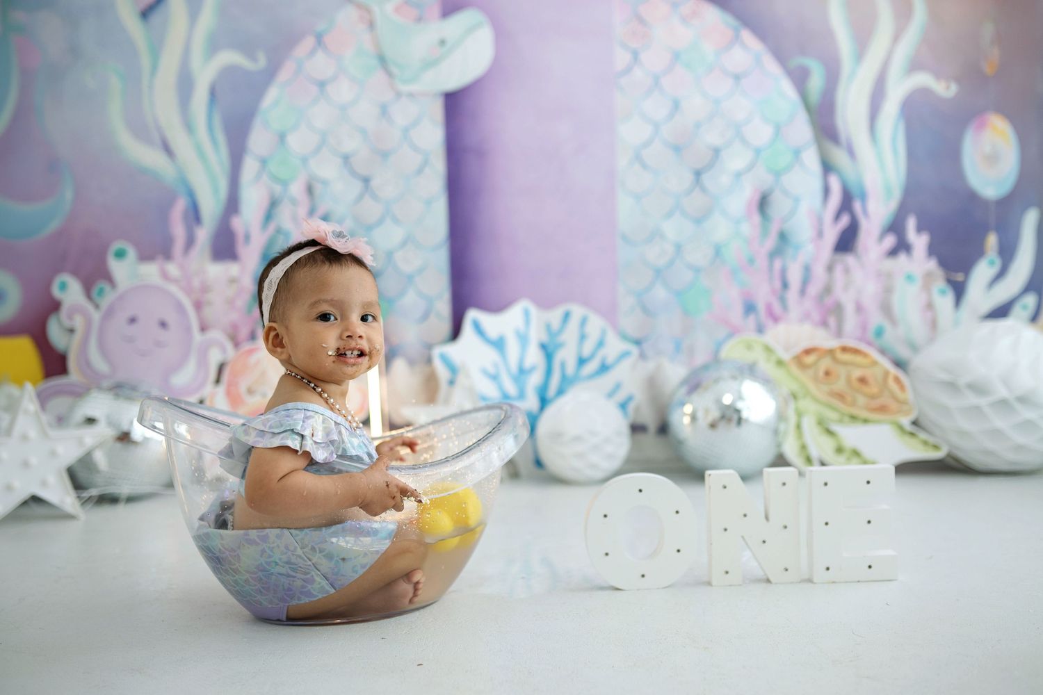 Baby in blue outfit sits on white floor with magical underwater themed backdrop and decorative elements.