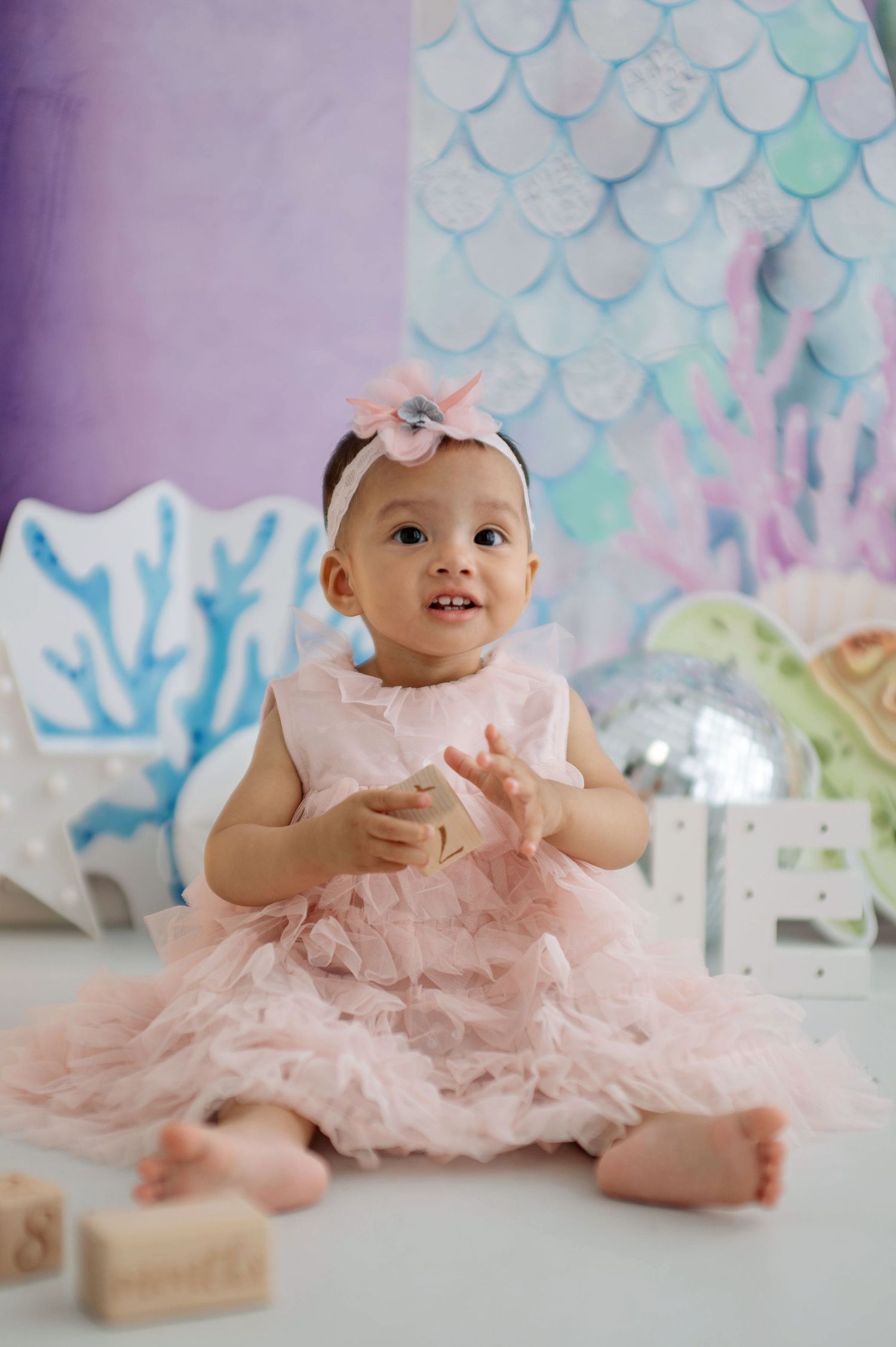 Sweet baby wearing pink tulle dress sits on floor against pastel backdrop during cake smash photo session.