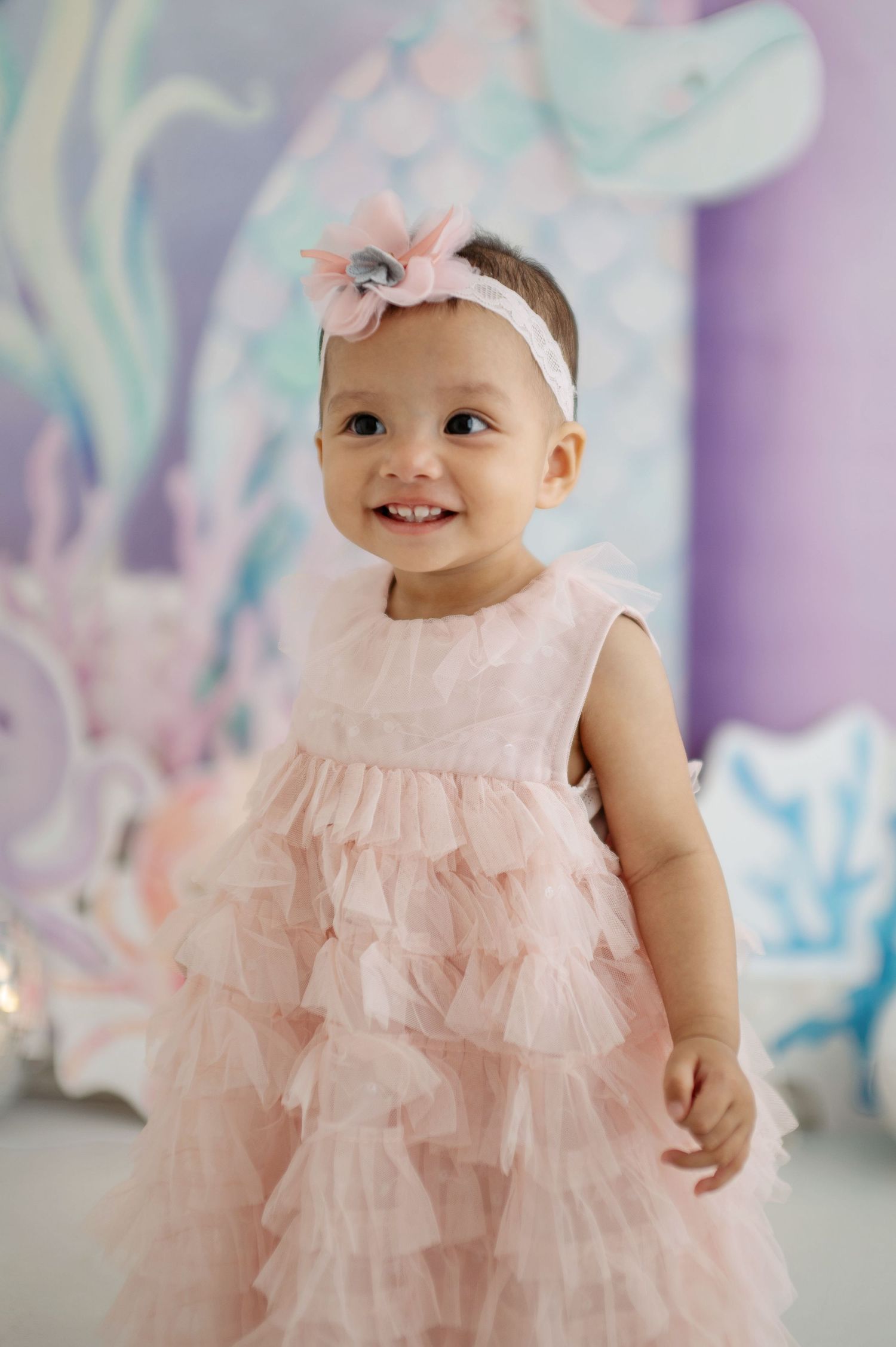 Baby in delicate pink dress and white headband poses against dreamy pastel background.