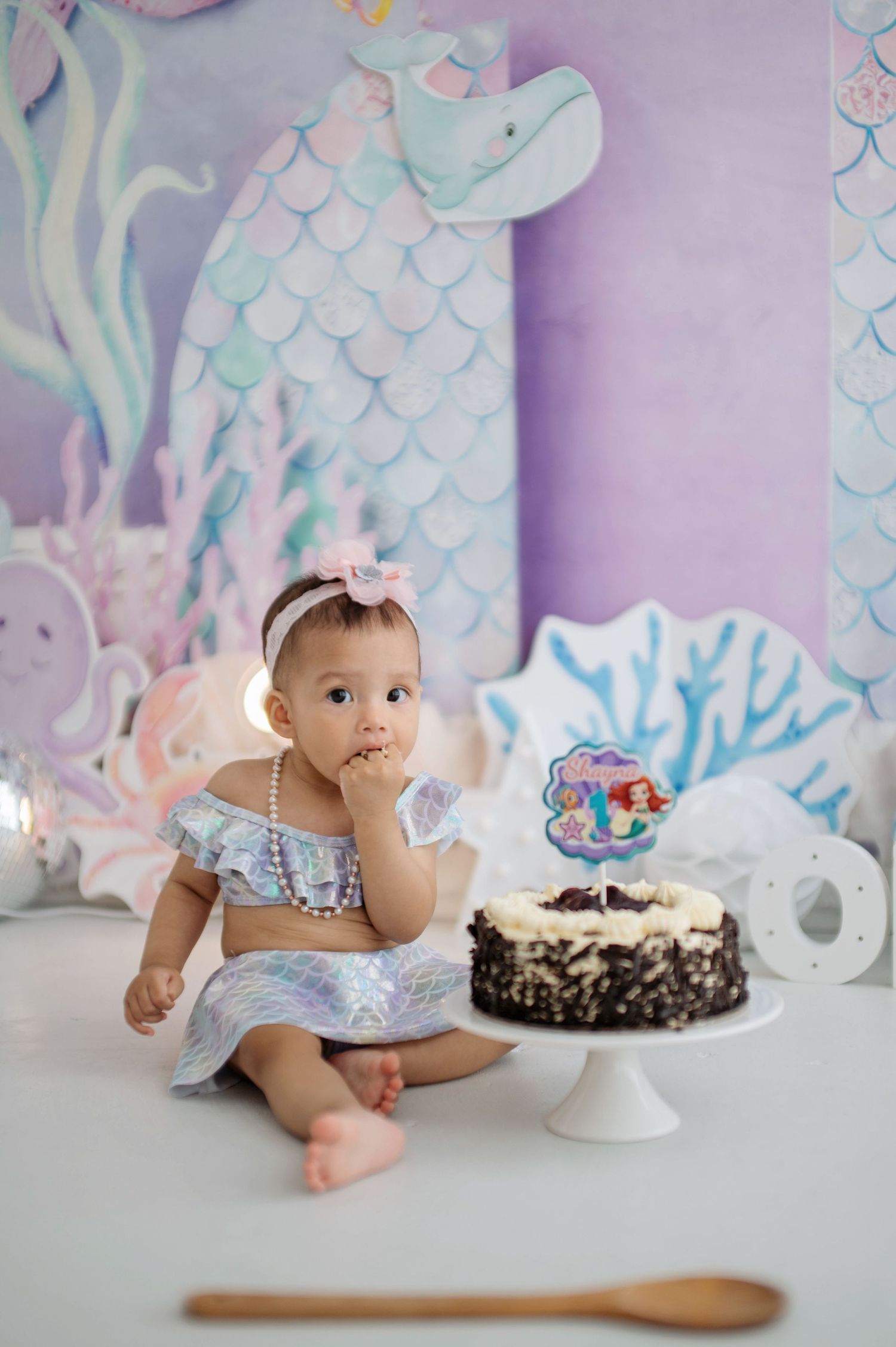 Baby in blue outfit crawls towards birthday cake on white cake stand against pastel mermaid-themed backdrop.