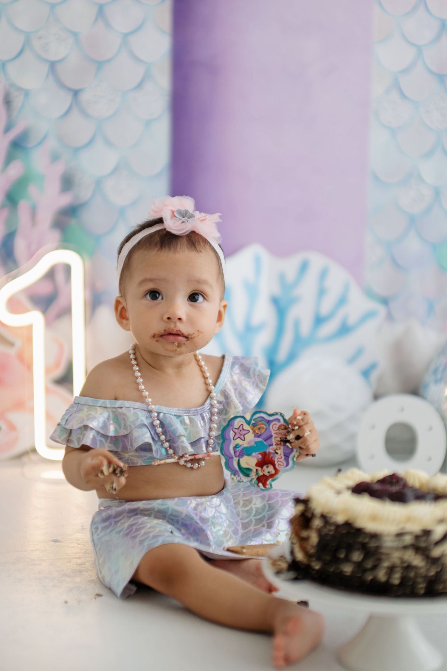 Baby in sparkly blue outfit sits on white fur with mermaid-themed backdrop and cake smash setup.
