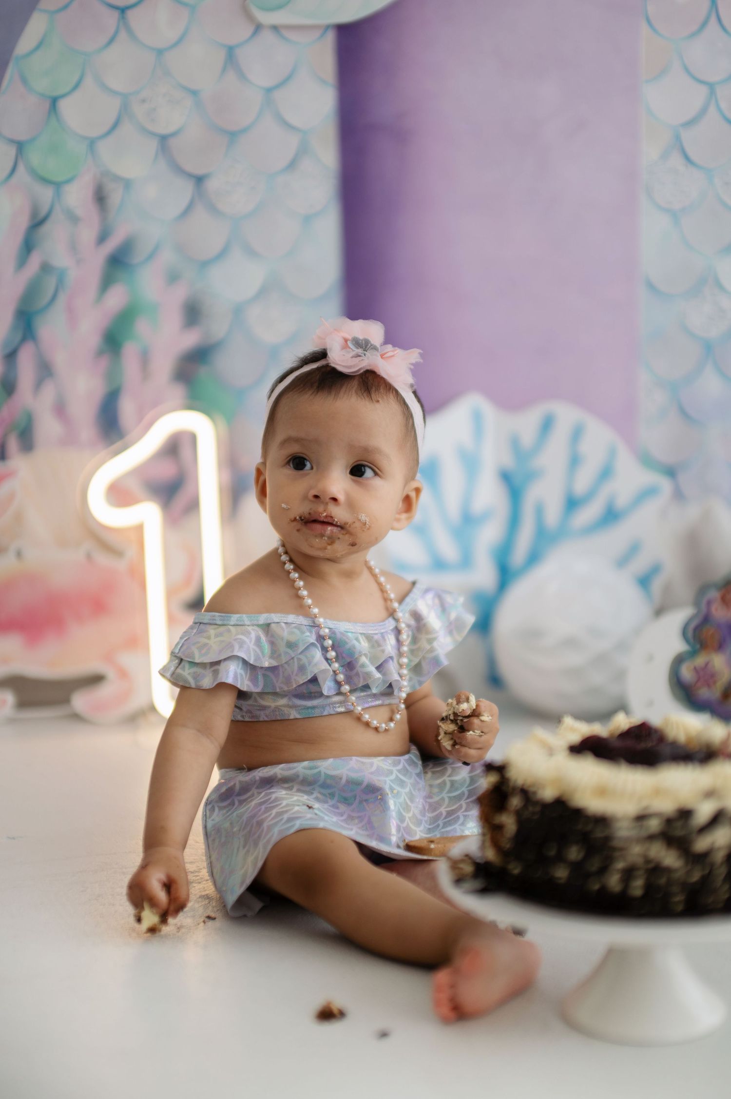Baby in blue ruffled outfit sits on floor next to smashed birthday cake during first birthday cake smash photo session.