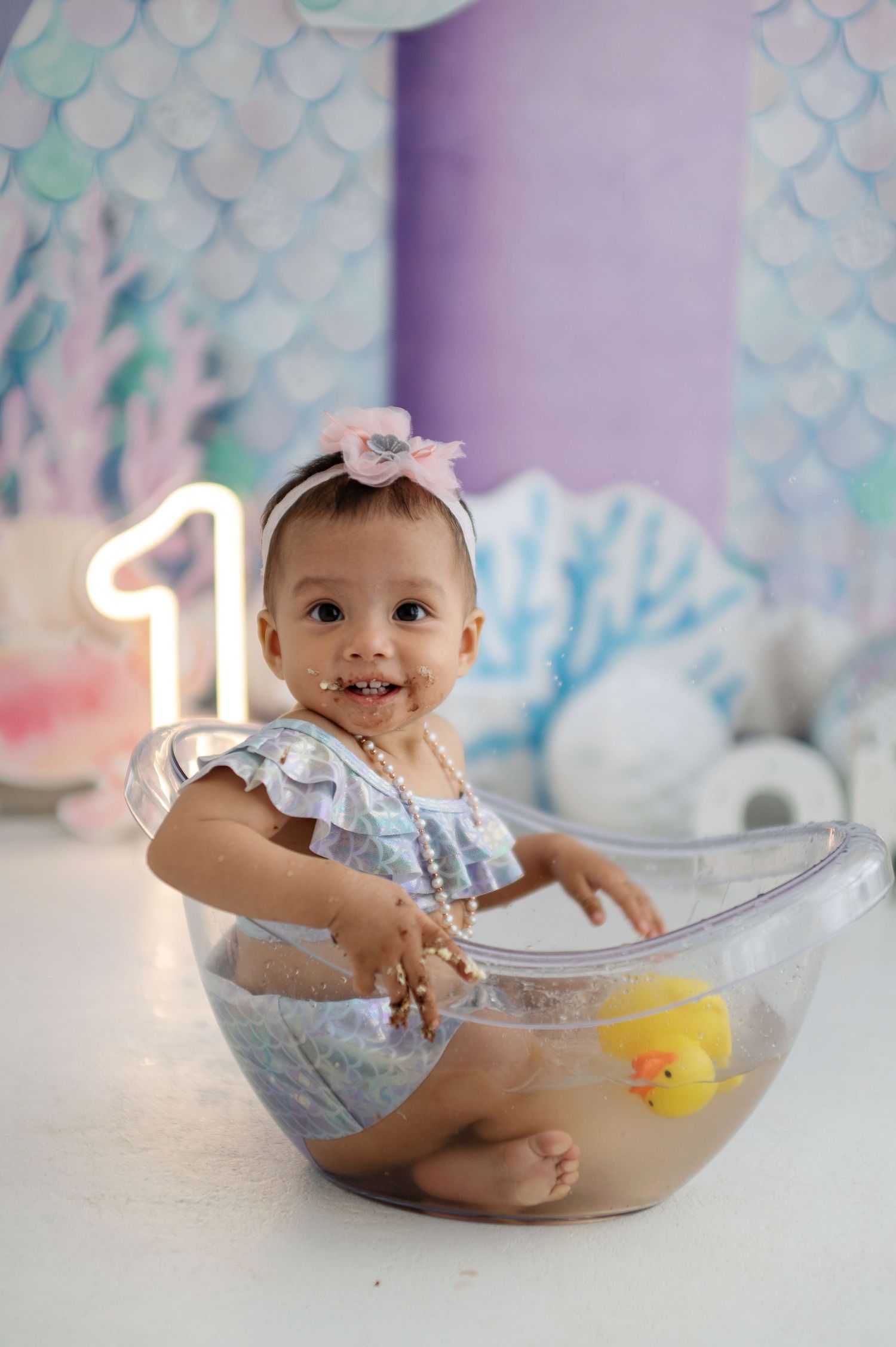 Baby plays with cake during first birthday photoshoot with mermaid-themed decorations in background.