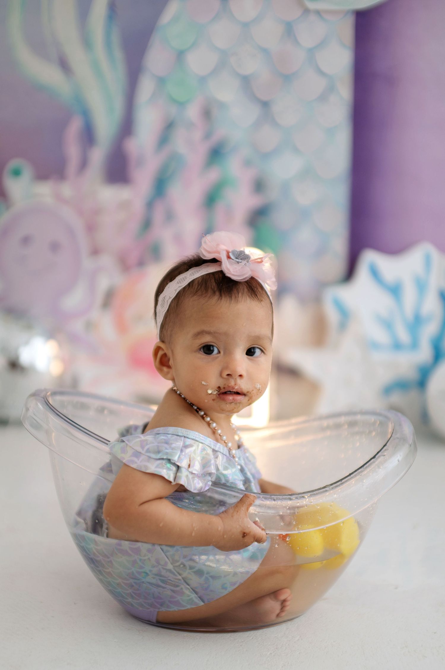 Baby sits in clear bowl with cake during first birthday photo shoot against purple and teal background.