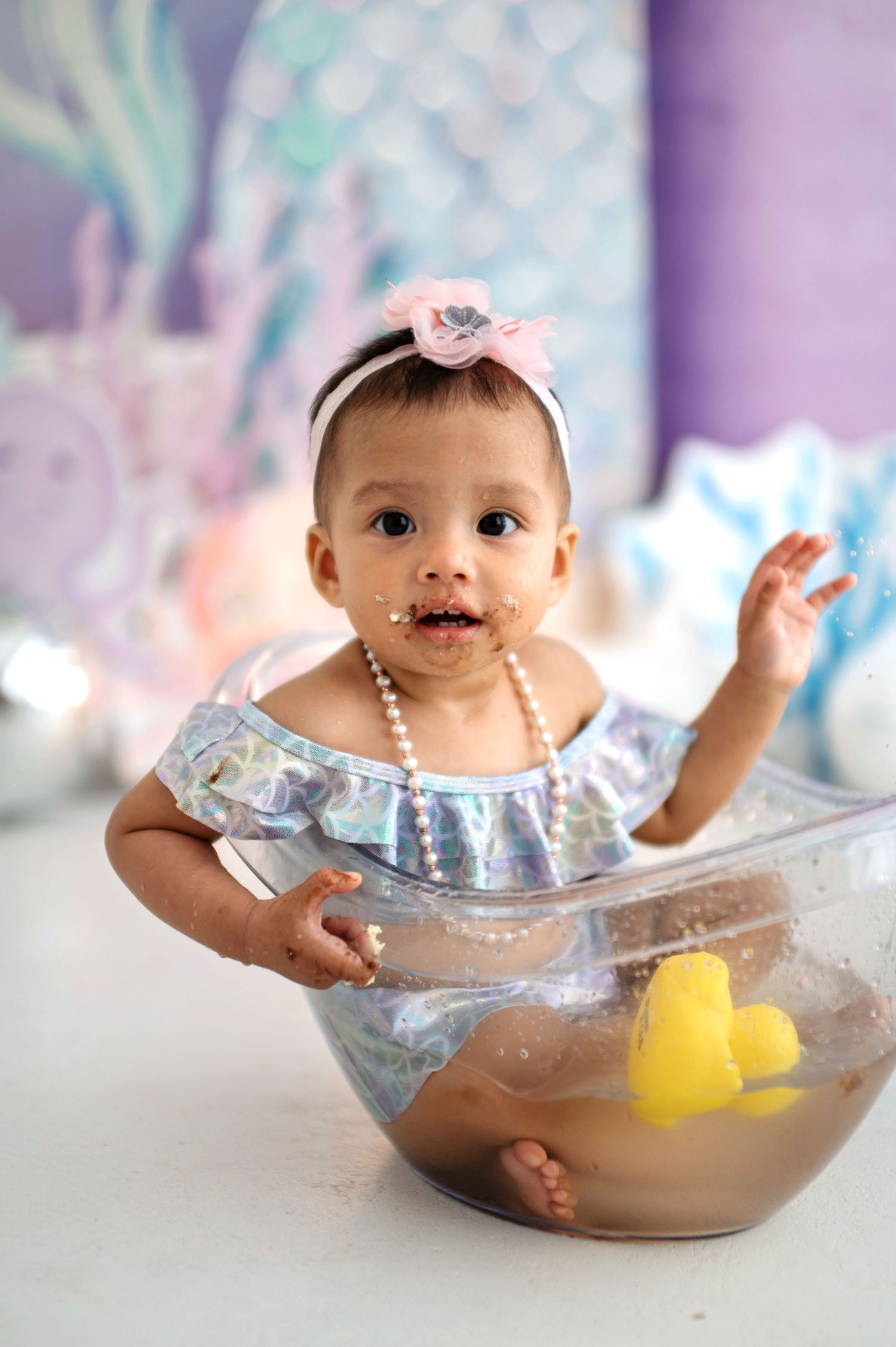 Baby sits in clear bowl with chocolate cake smeared on face during messy cake smash photo session.