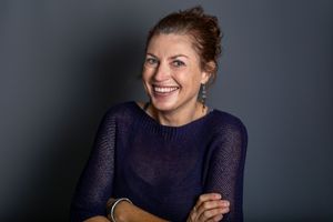 A person wearing a navy blue sweater laughs joyfully while posing against a gray background.