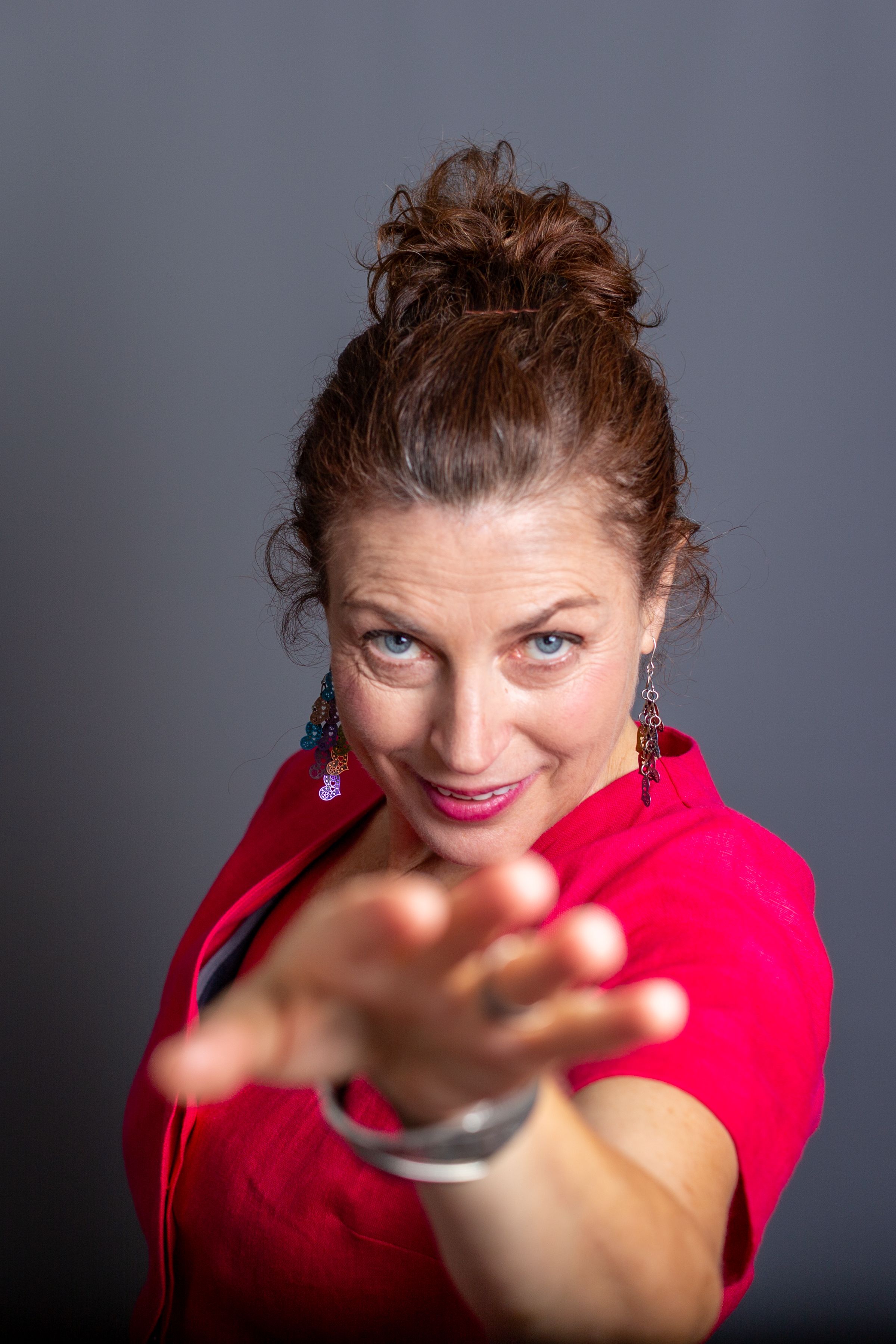A person in a bright red top reaches toward the camera with an engaging expression against a gray background.