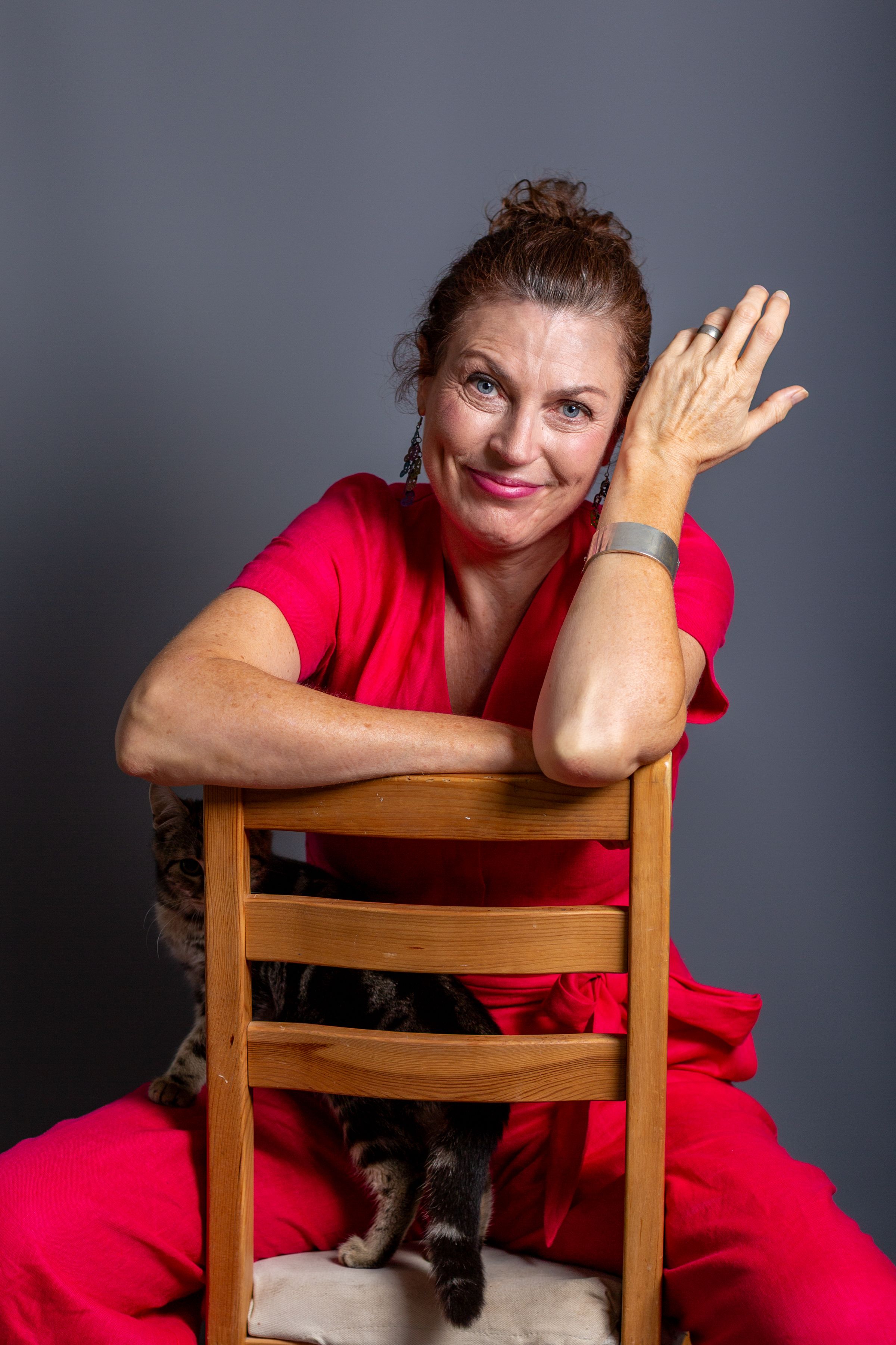 A person in a bright red outfit poses playfully on a wooden chair against a dark background.