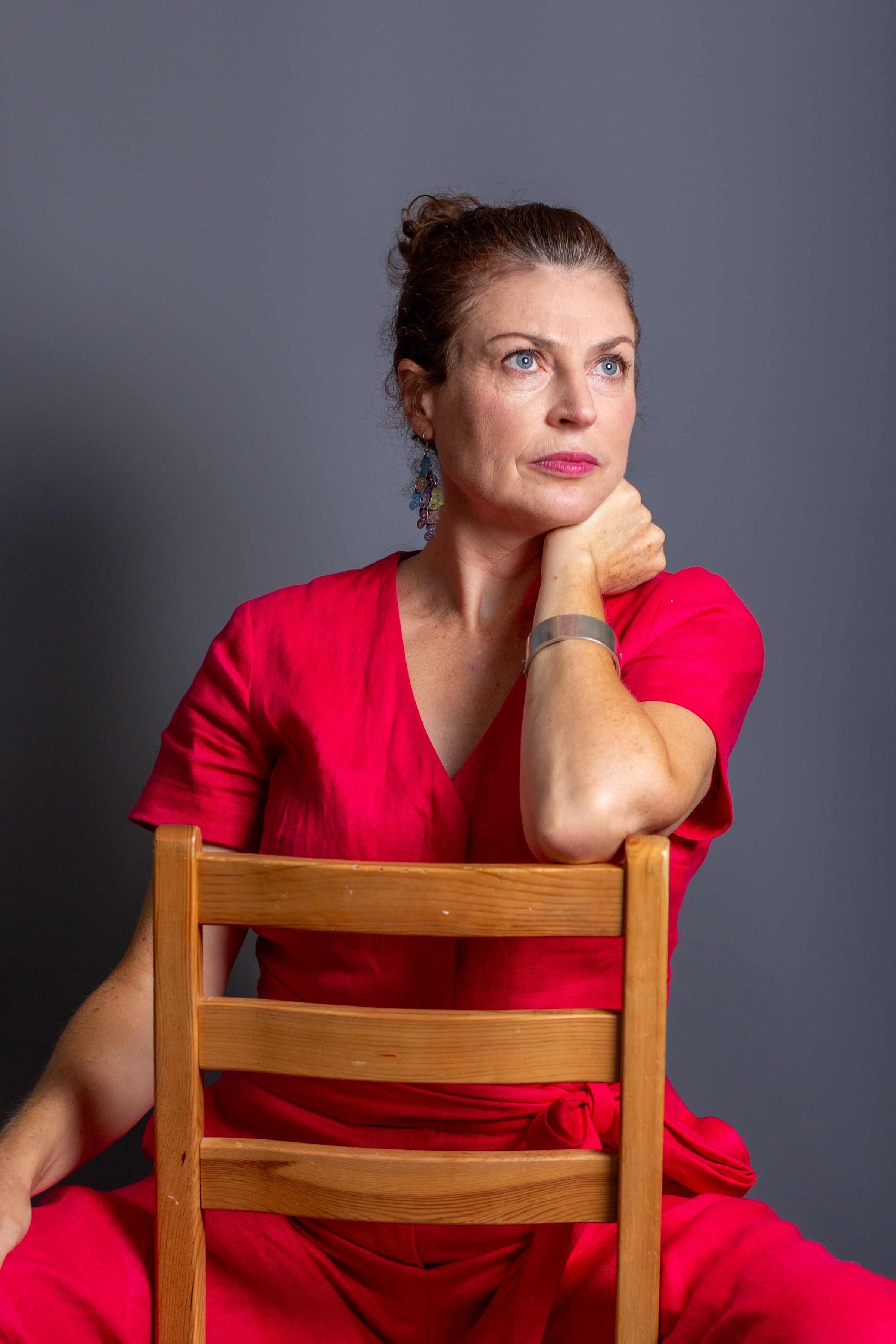 A person in a vibrant red outfit sits thoughtfully on a wooden chair against a gray backdrop.