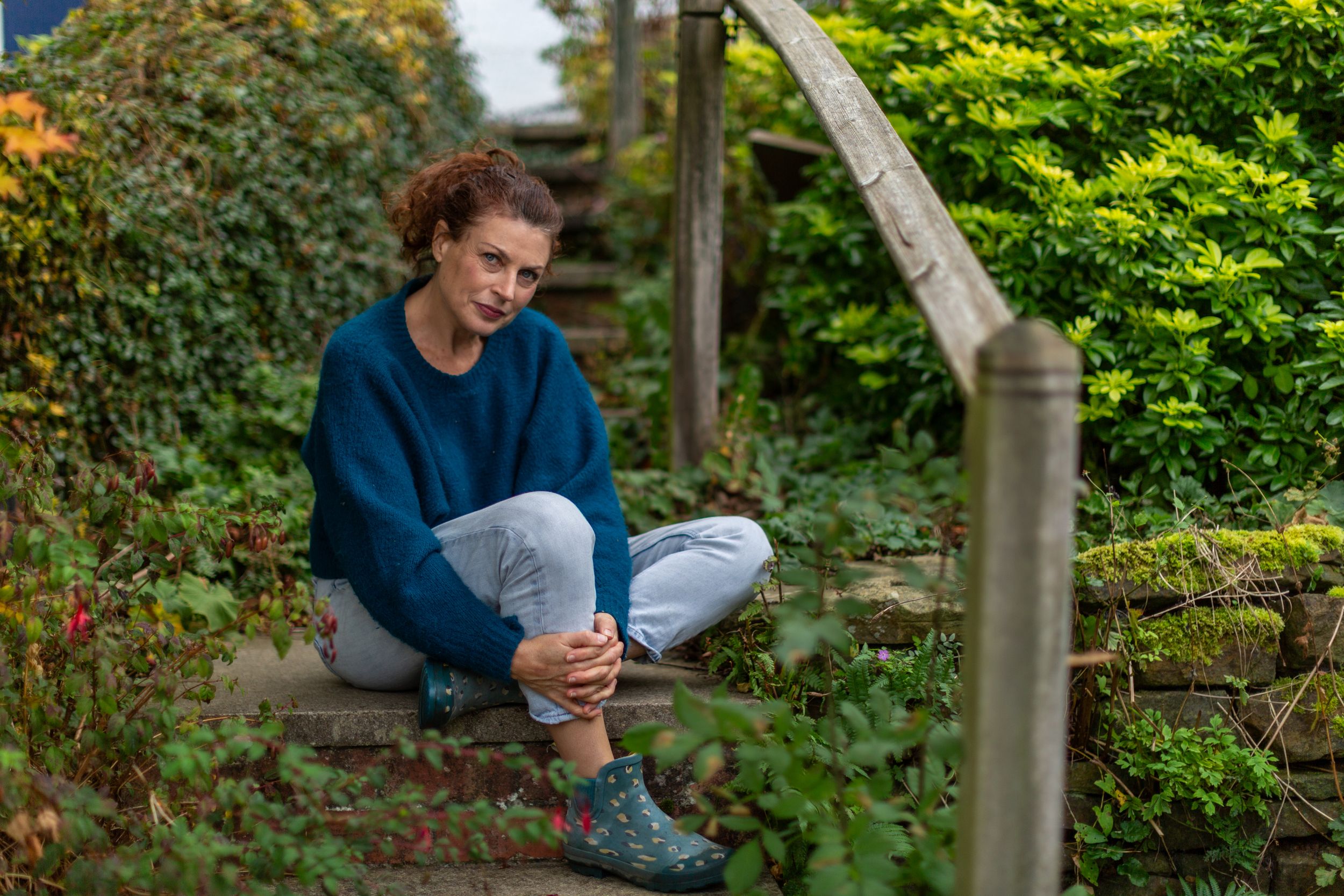 A person sitting on a wooden bench in a lush garden setting wearing casual blue clothing.