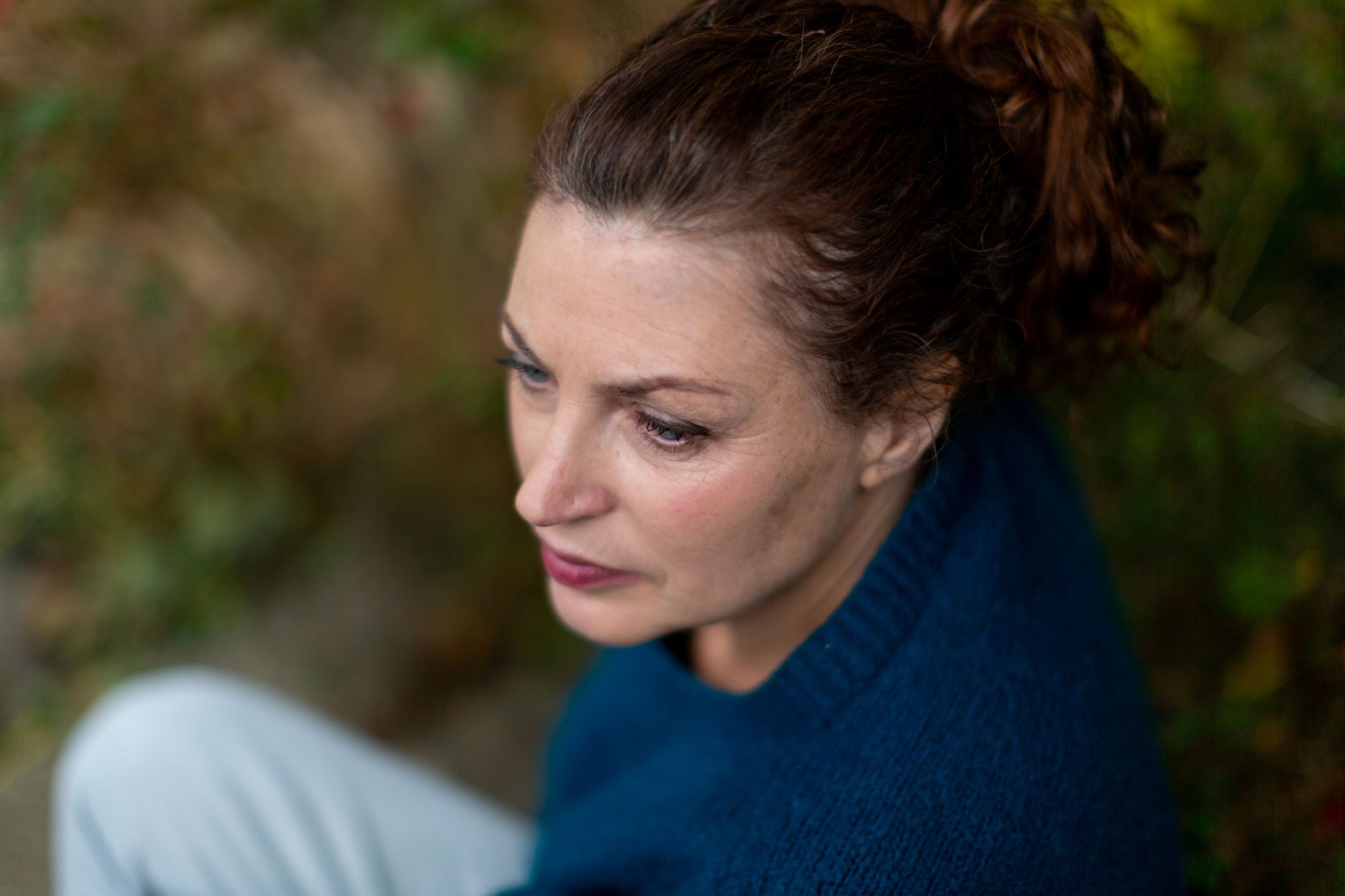 Profile view of someone wearing a blue sweater with their hair in a bun against a blurred natural background.