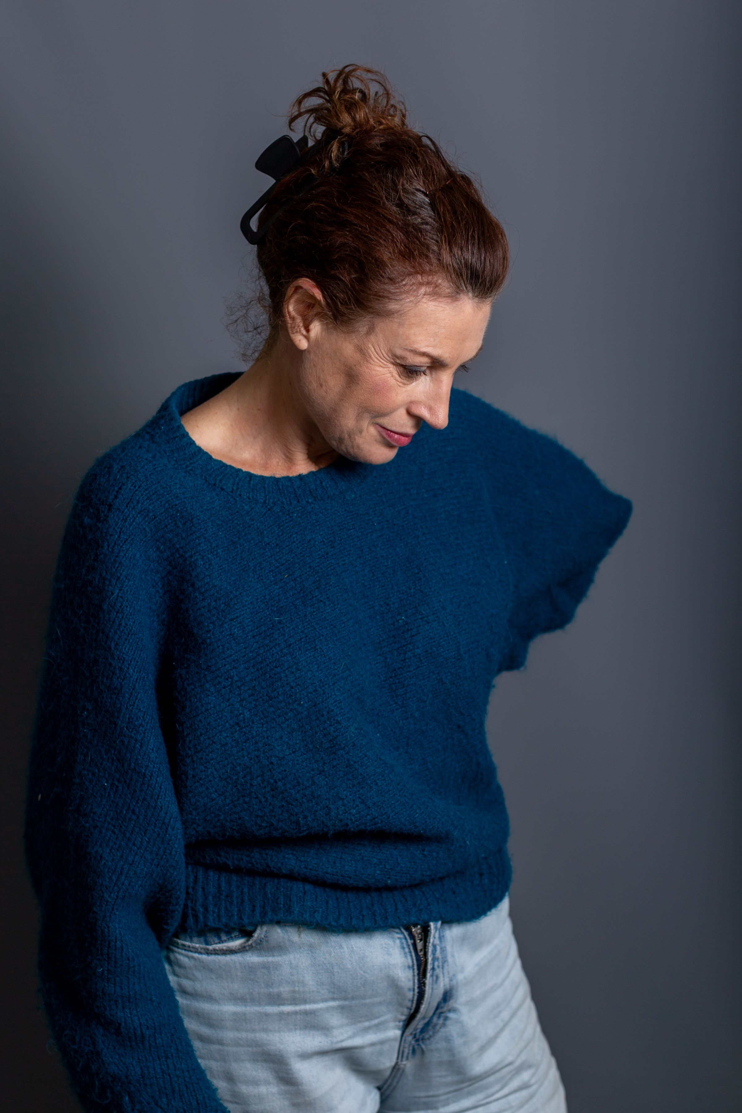 A person wearing a cozy navy blue sweater and light denim jeans poses against a dark gray background.