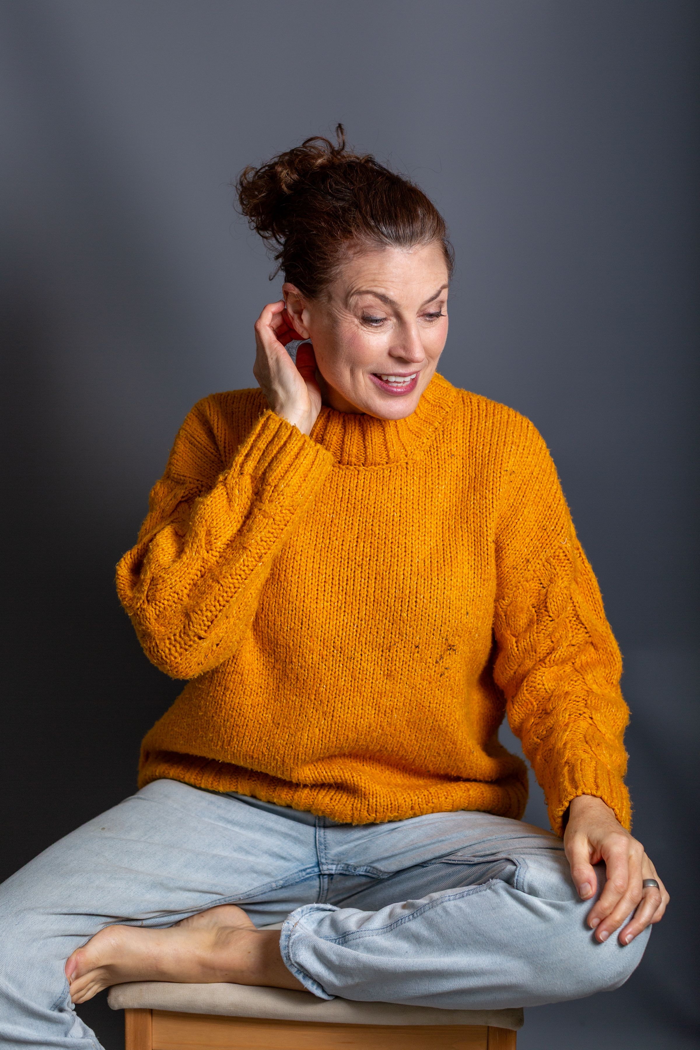 A person in a bright orange knit sweater sits cross-legged wearing light jeans against a gray backdrop.