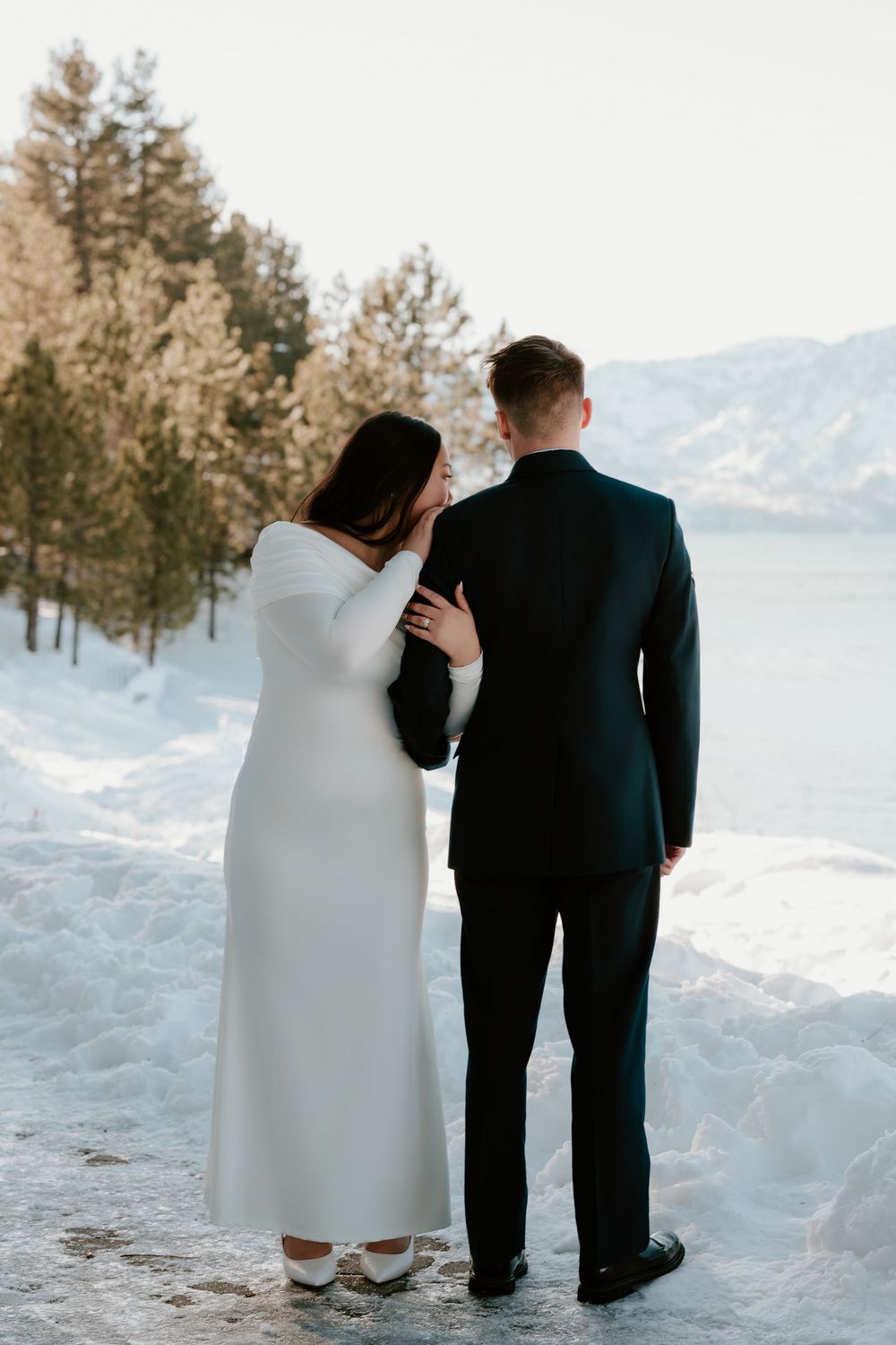 A couple in formal wedding attire stands together in snowy scenery with a mountain lake view in the background.