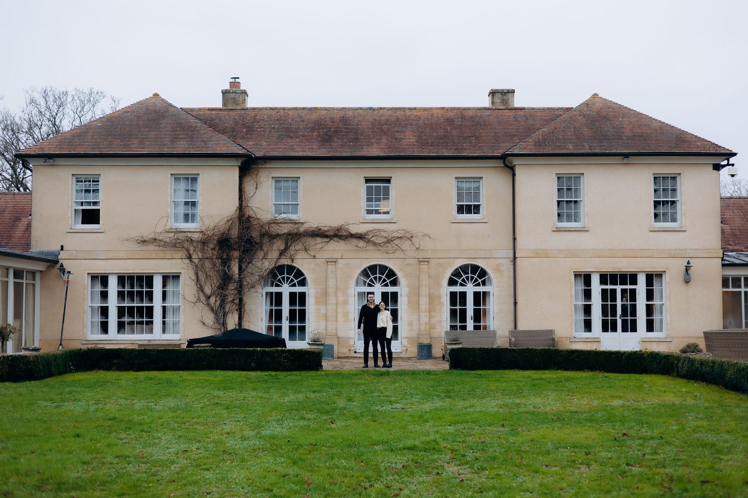 Elegant cream-colored mansion with bay windows and manicured hedges features two figures standing in the arched doorway.