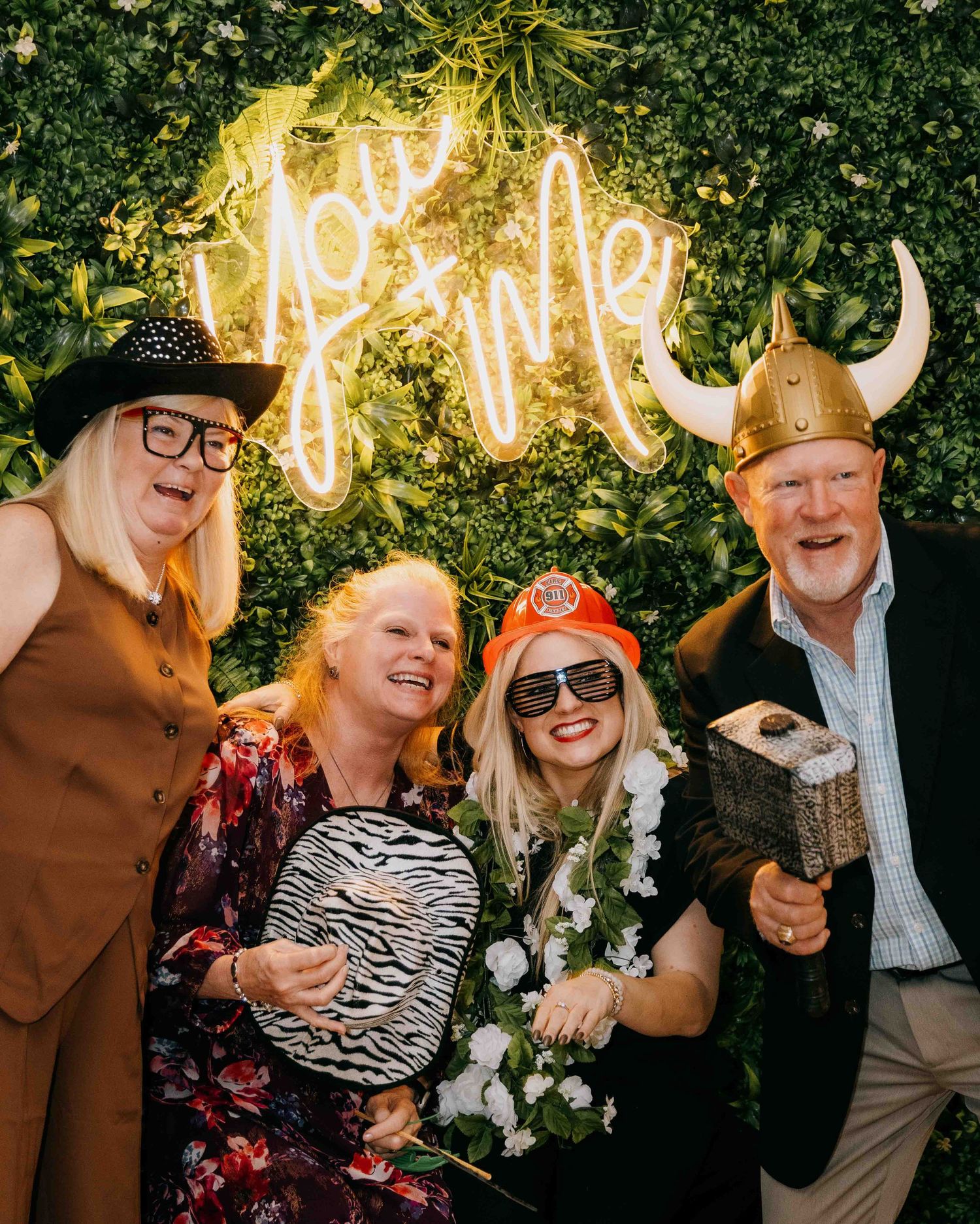 Group of friends strike silly poses and wear props for a wedding photo booth against a green hedge wall and neon sign in the background.