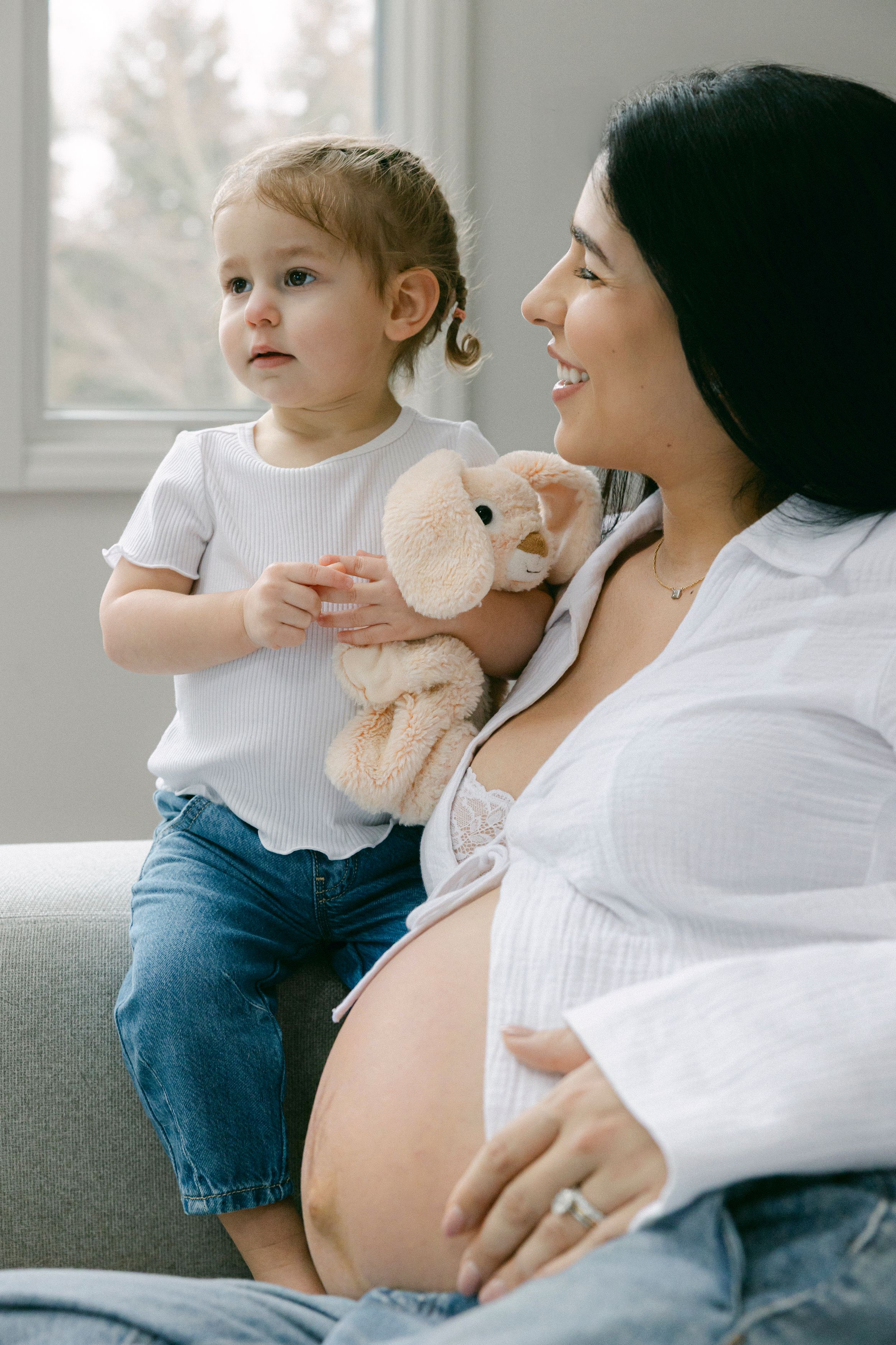 Expectant mother in white shirt sits on couch with toddler holding stuffed animal during intimate family moment.