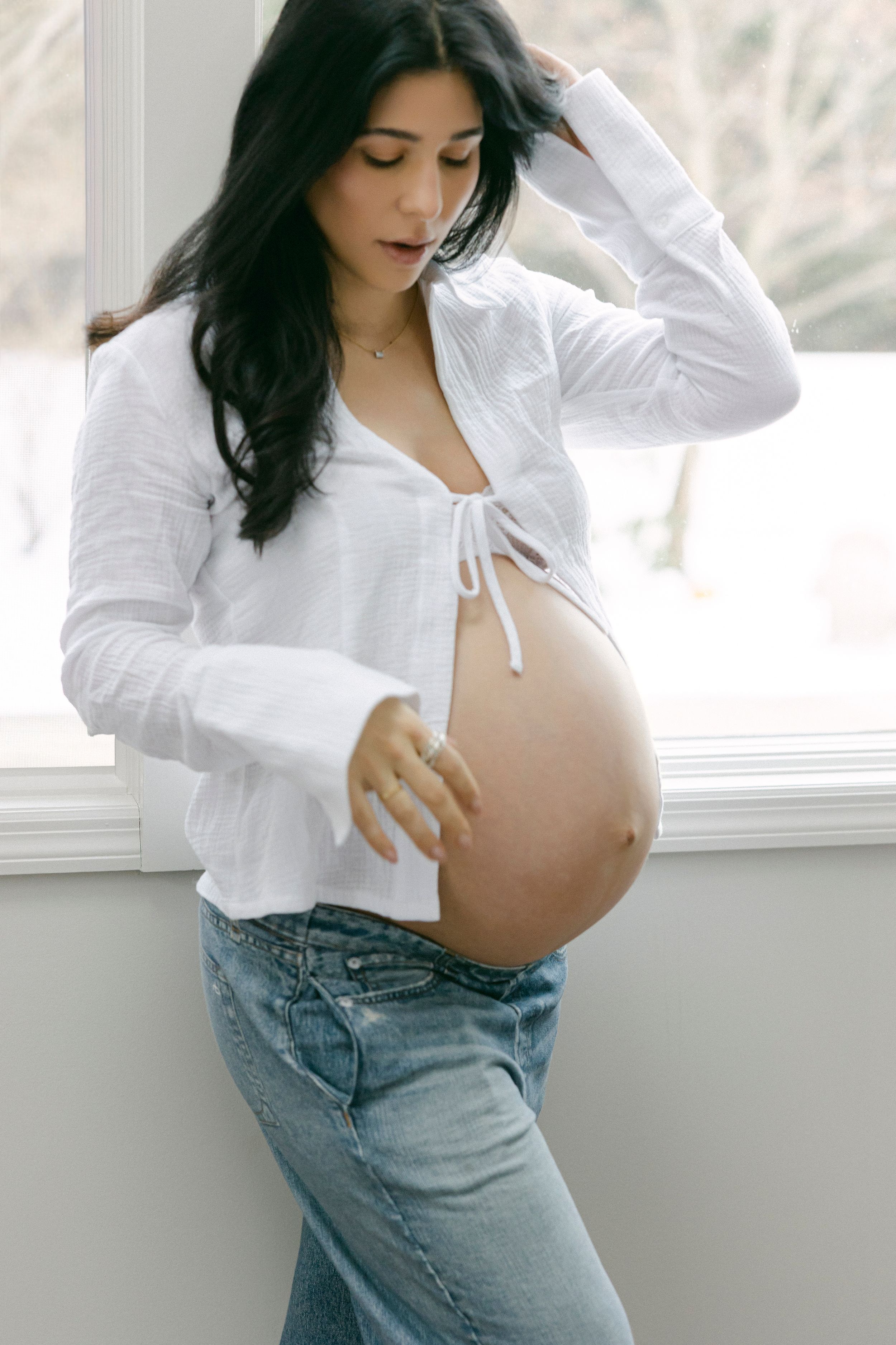 A person in a white button-down shirt and jeans poses against a bright window in a casual lifestyle photoshoot.