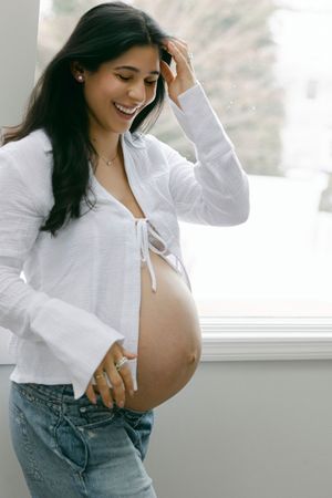 A pregnant woman in a white blouse and jeans stands by a window, smiling and touching her baby bump.