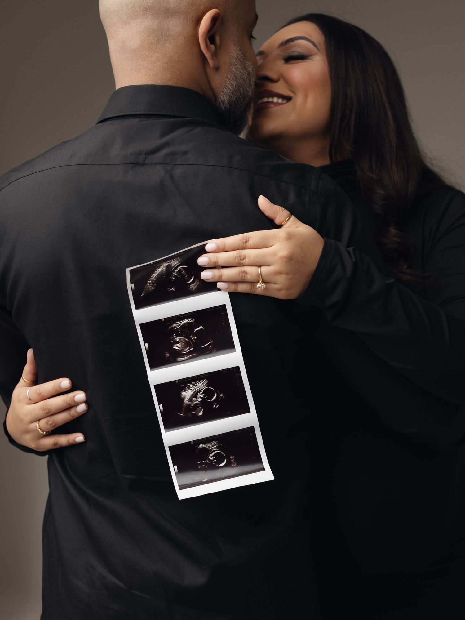 A couple embraces while holding ultrasound photos, celebrating their pregnancy announcement in a moody artistic style. Elizabeth Lang Photog