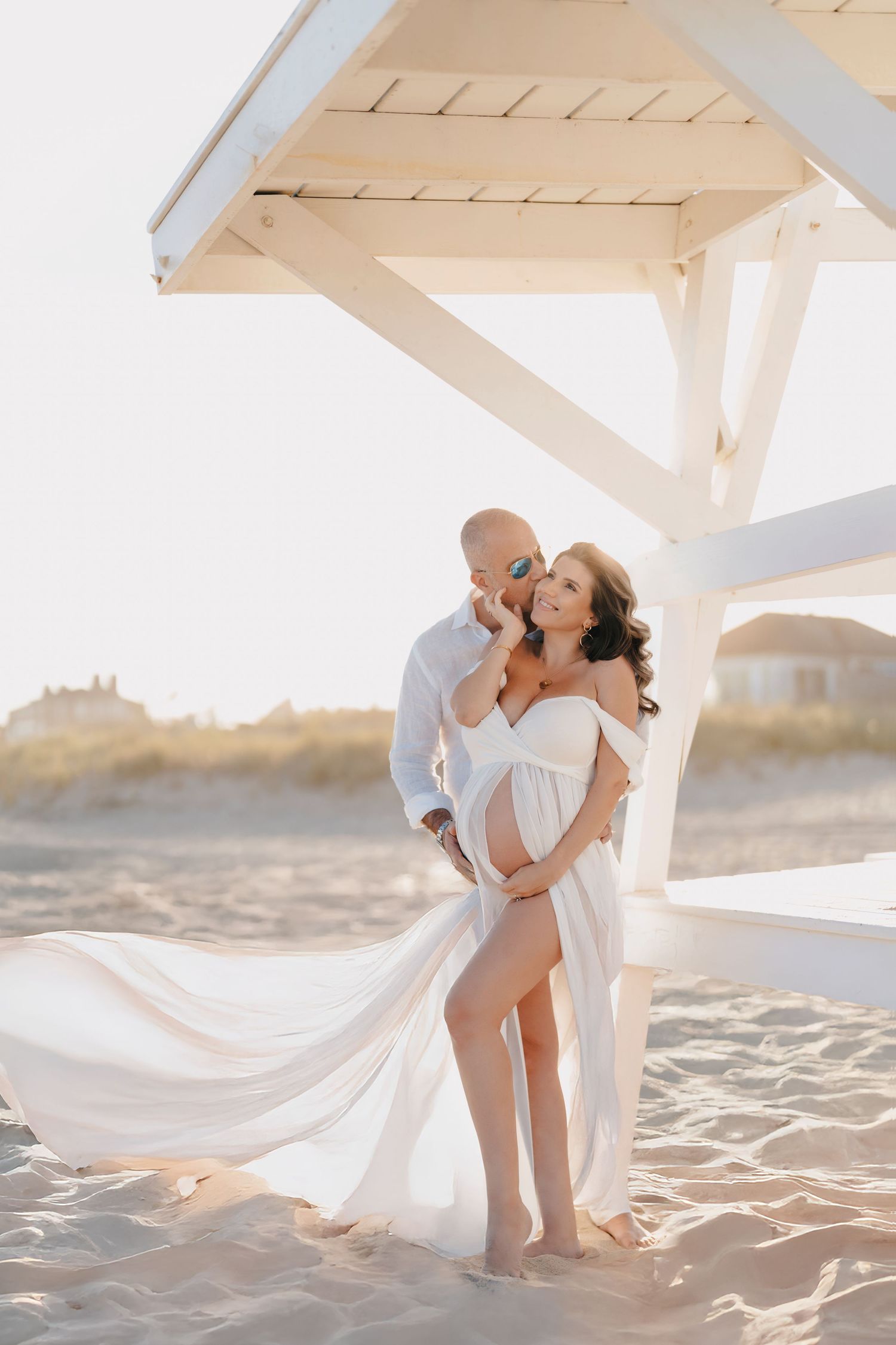 Romantic couple embraces on sandy beach at sunset under white wooden gazebo with flowing white dress.
