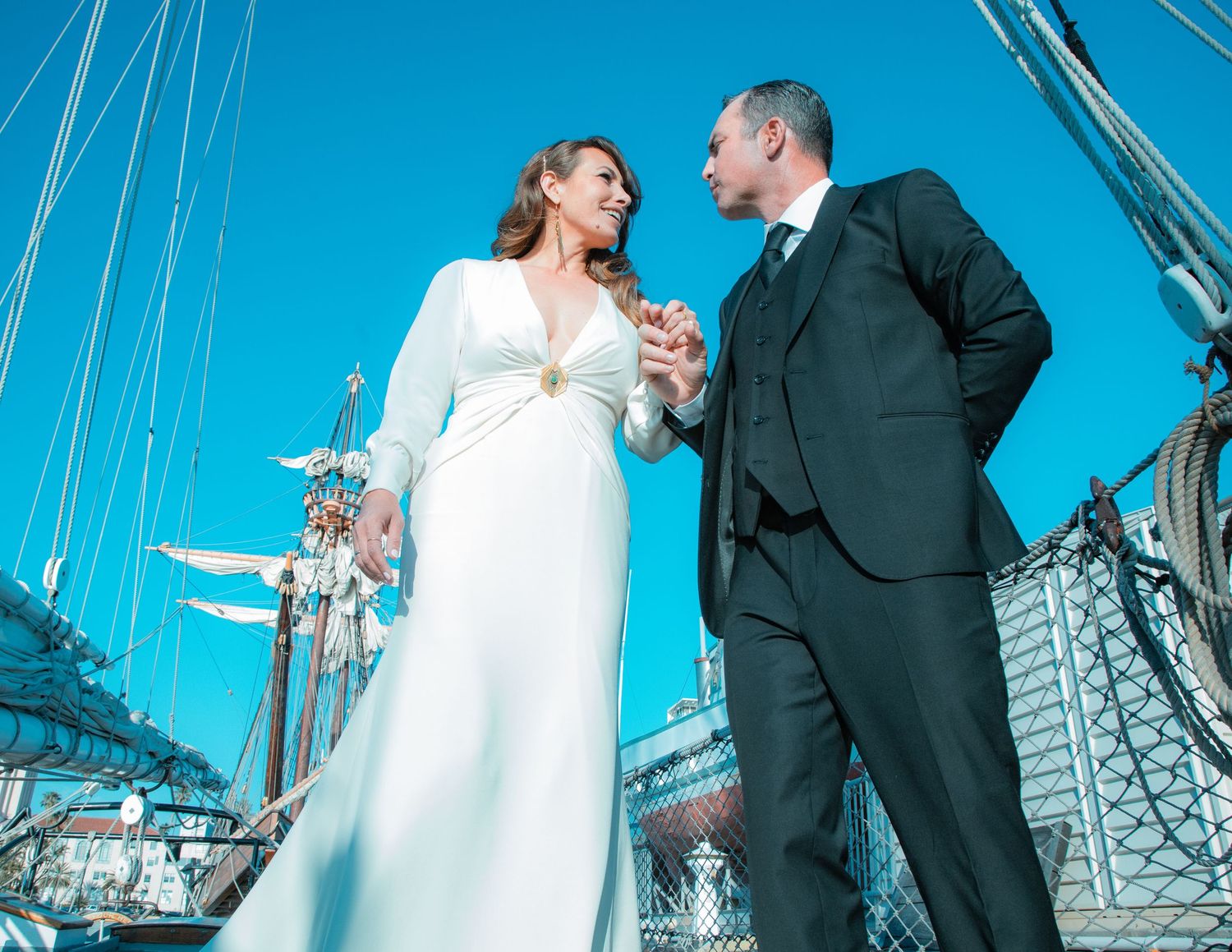 A person in a floral dress officiates a wedding ceremony on a boat deck as a couple embraces.