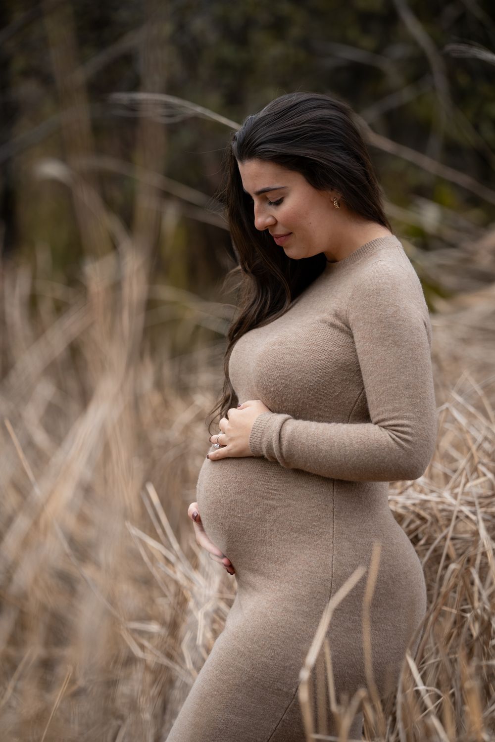 A pregnant woman in a beige sweater poses in a field of dried grass during a maternity photo session.