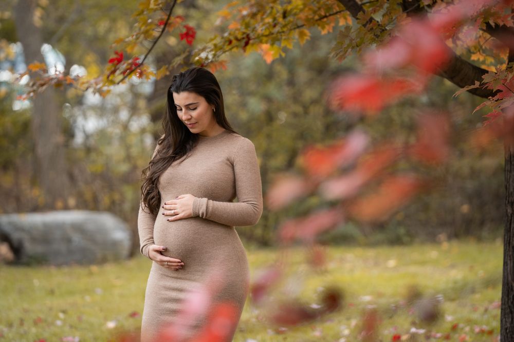 Expectant mother in beige sweater dress poses in autumn garden with colorful fall foliage in background.