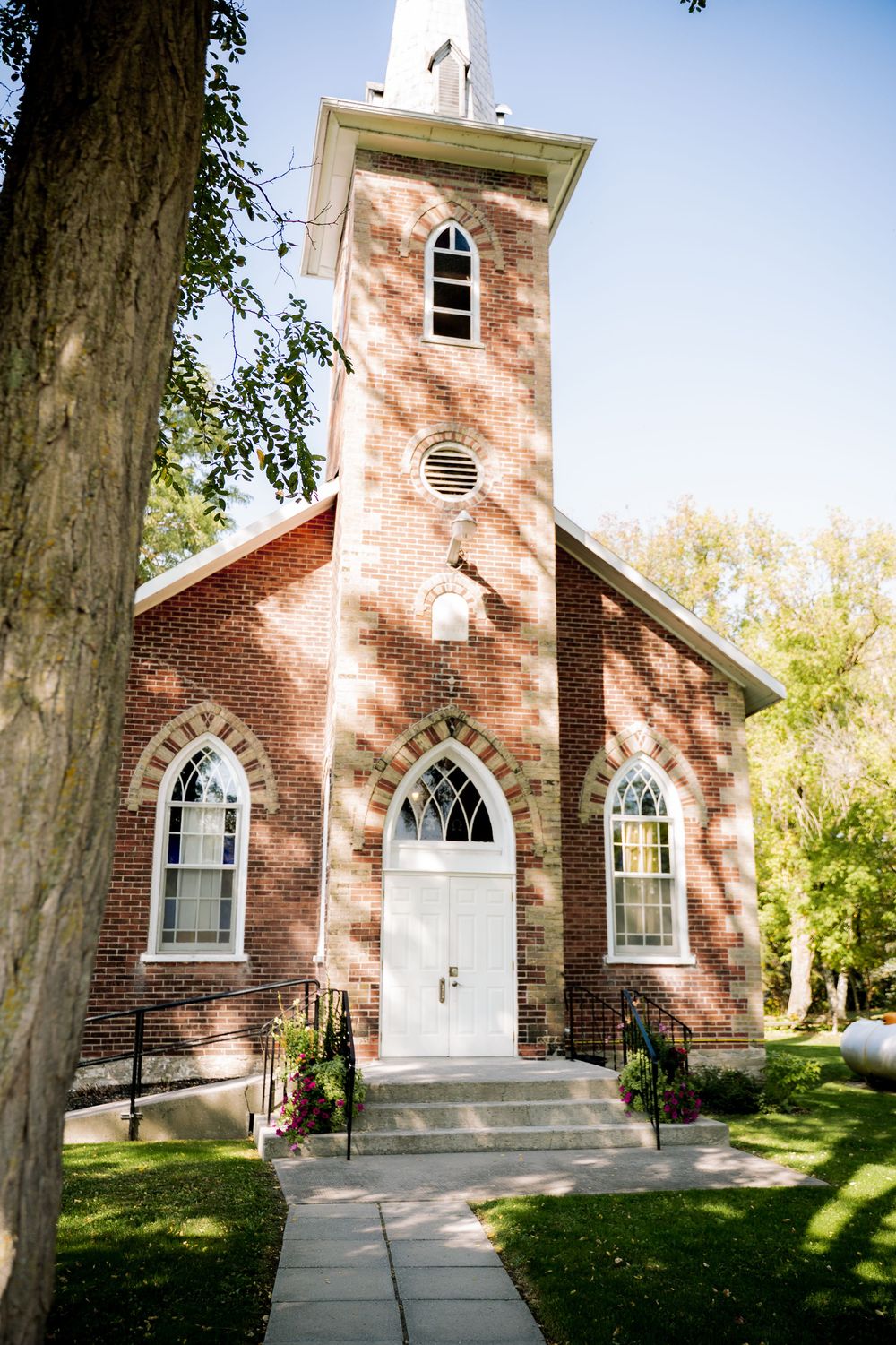 Historic red brick church with Gothic arched windows and white trim features a tall steeple and welcoming entrance.