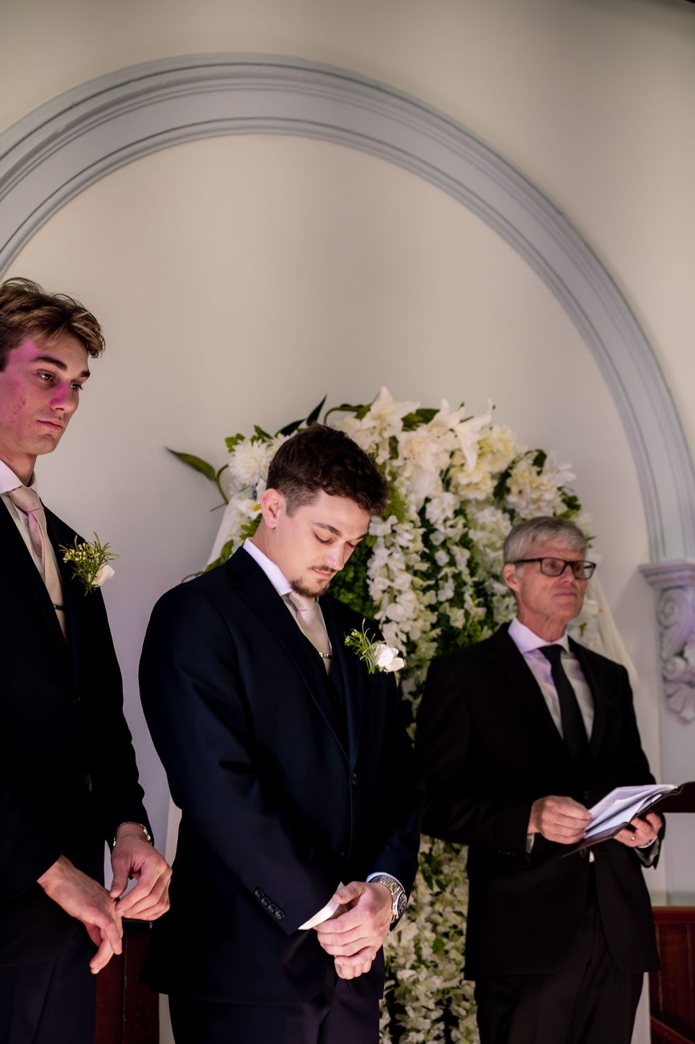 Wedding ceremony participants stand solemnly under an elegant archway decorated with white floral arrangements.