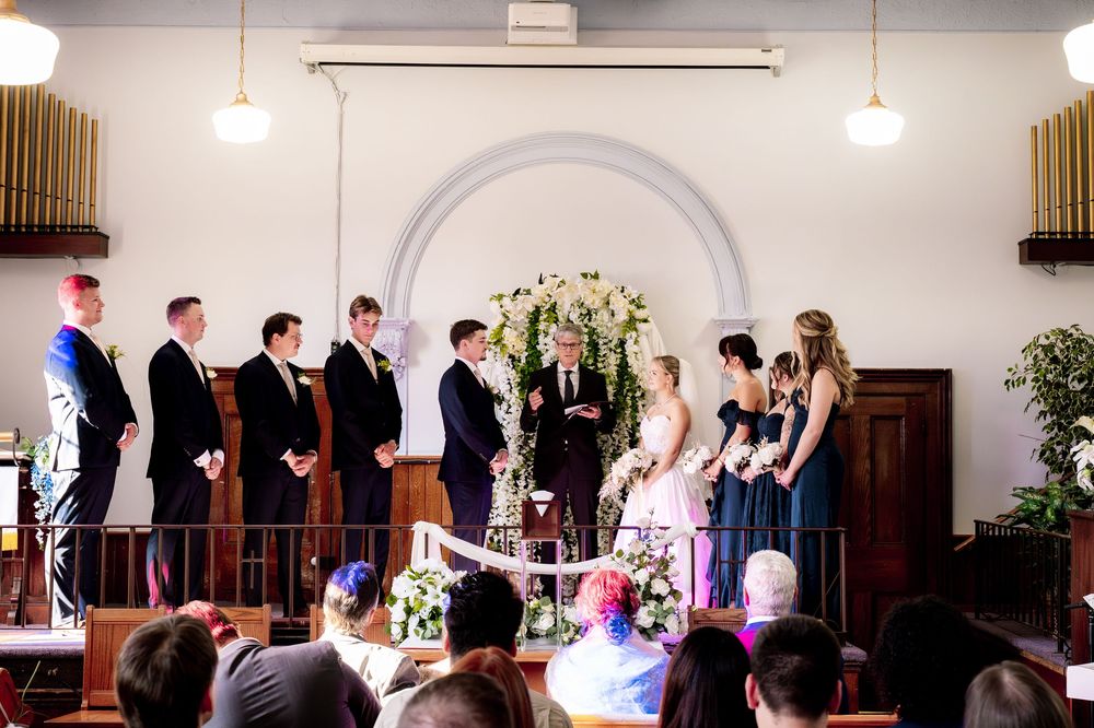 Wedding party stands in line at the altar during a traditional church ceremony with white floral decorations.